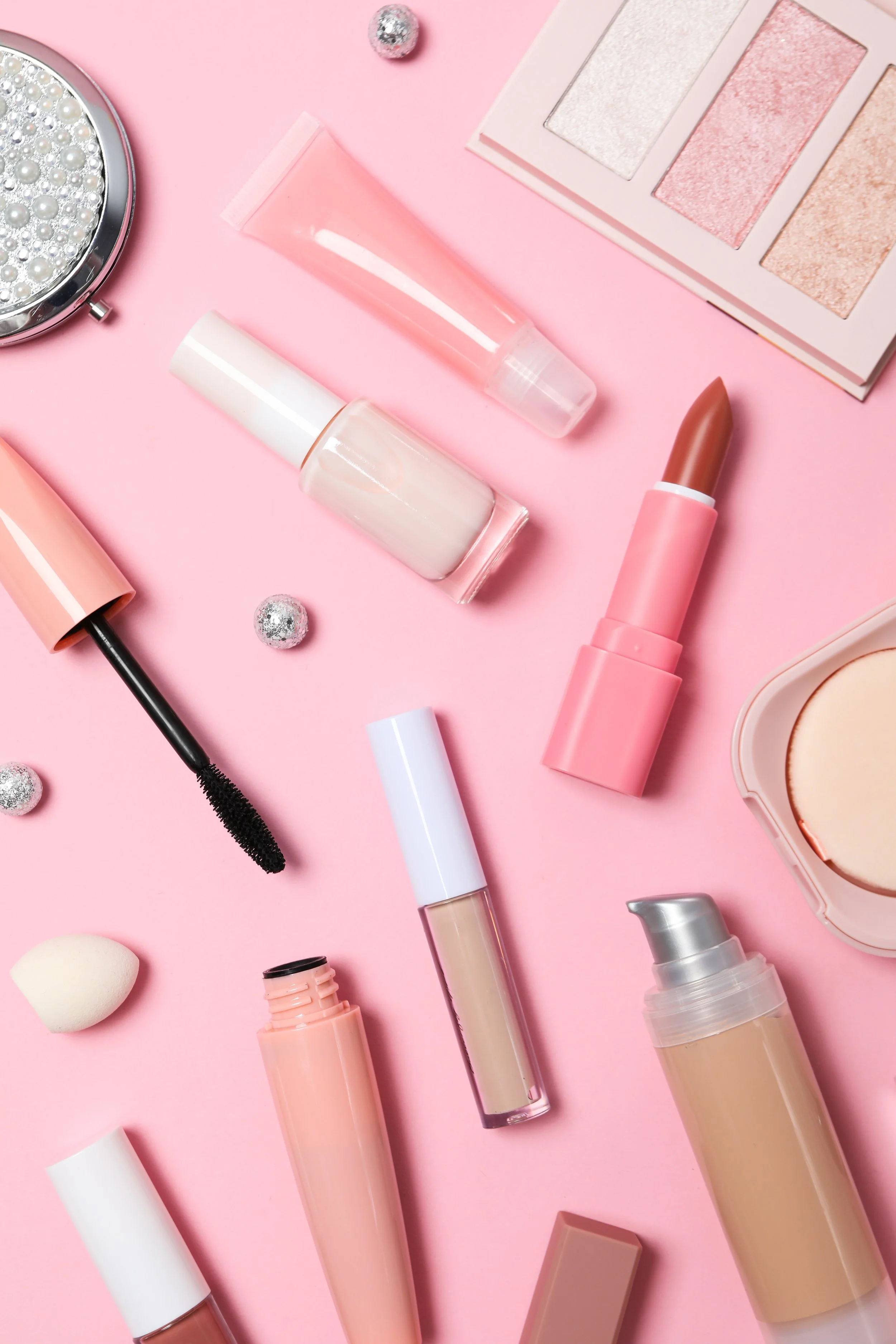 Arrangement of various makeup products, including lip balm, mascara, foundation, eyeshadow palette, and brushes, on a pink background.