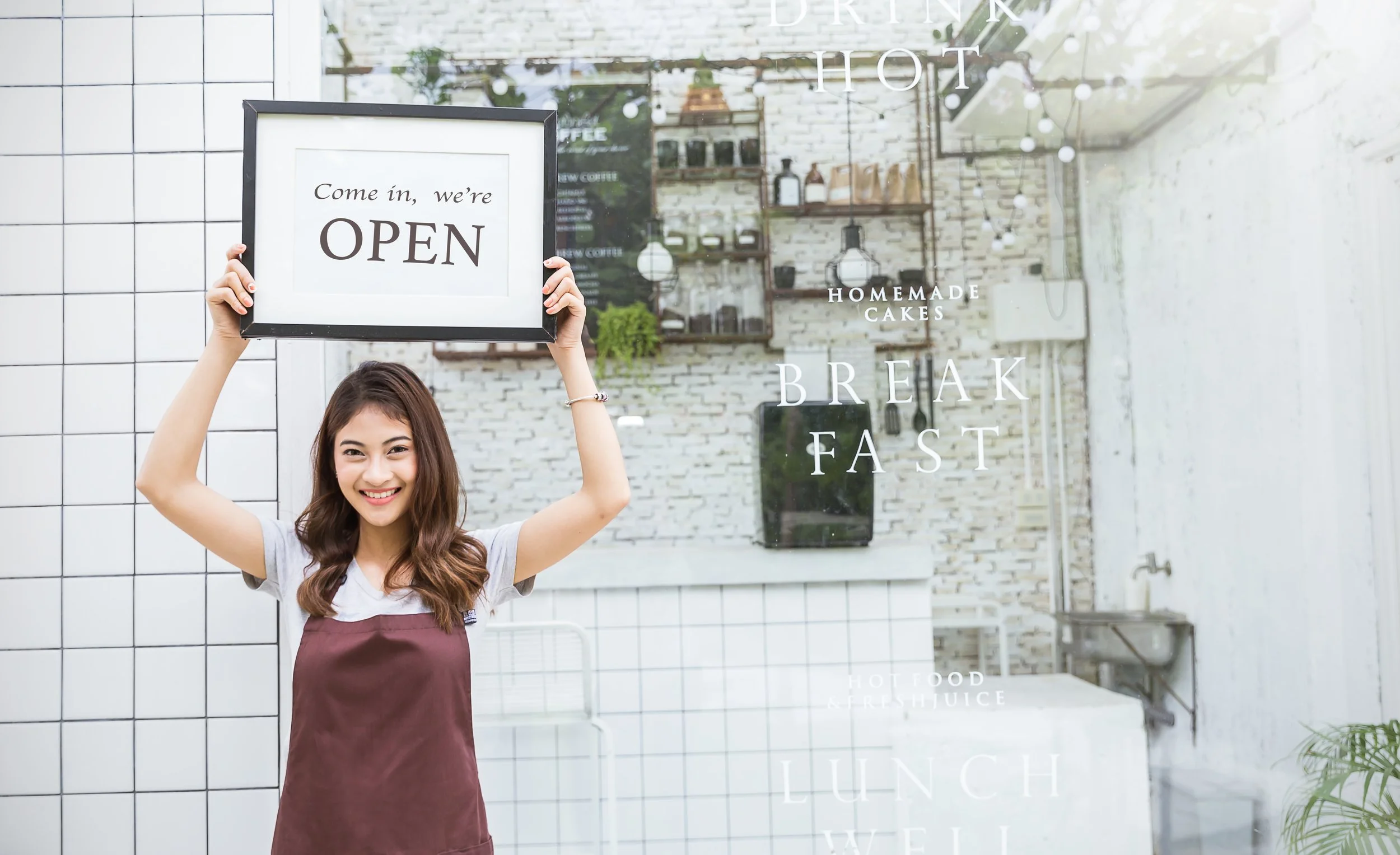 A young woman smiling and wearing an apron, holding a framed sign that says 'Come in, we're OPEN' in front of a cafe window with white text advertising homemade cakes, breakfast, lunch, and juice.