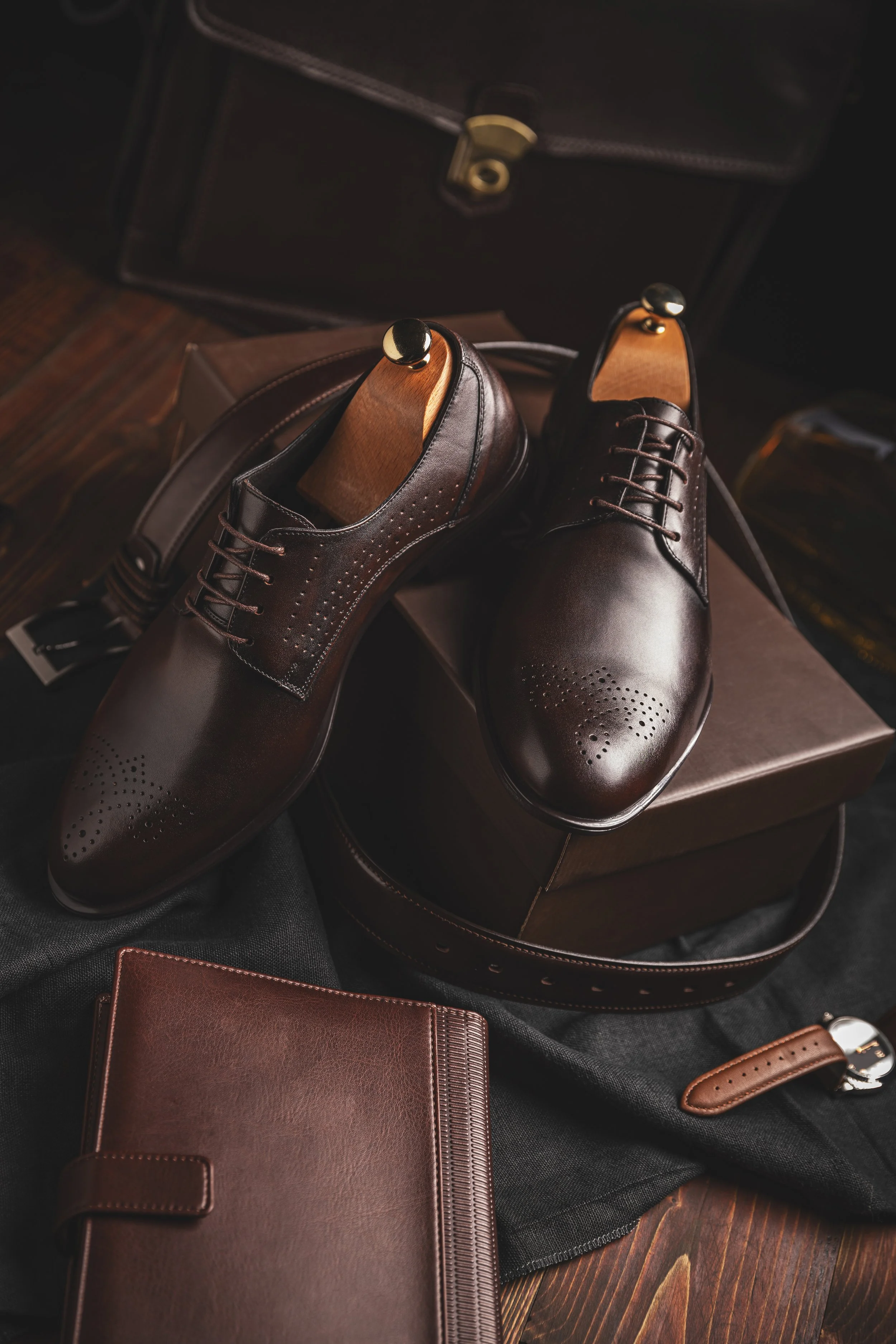 Pair of polished brown dress shoes with perforated details, placed on a brandy-colored box with accessories and a dark fabric underneath.