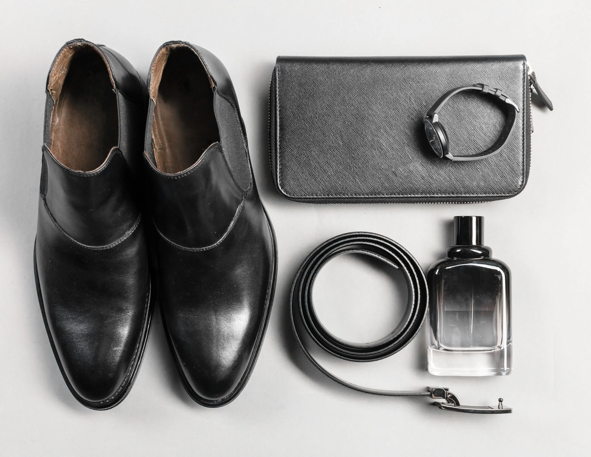 Pair of black leather dress shoes, a gray clutch purse with a black wristwatch on top, a black belt, a bottle of cologne or perfume, and a silver watch with a metal band, all arranged on a white surface.