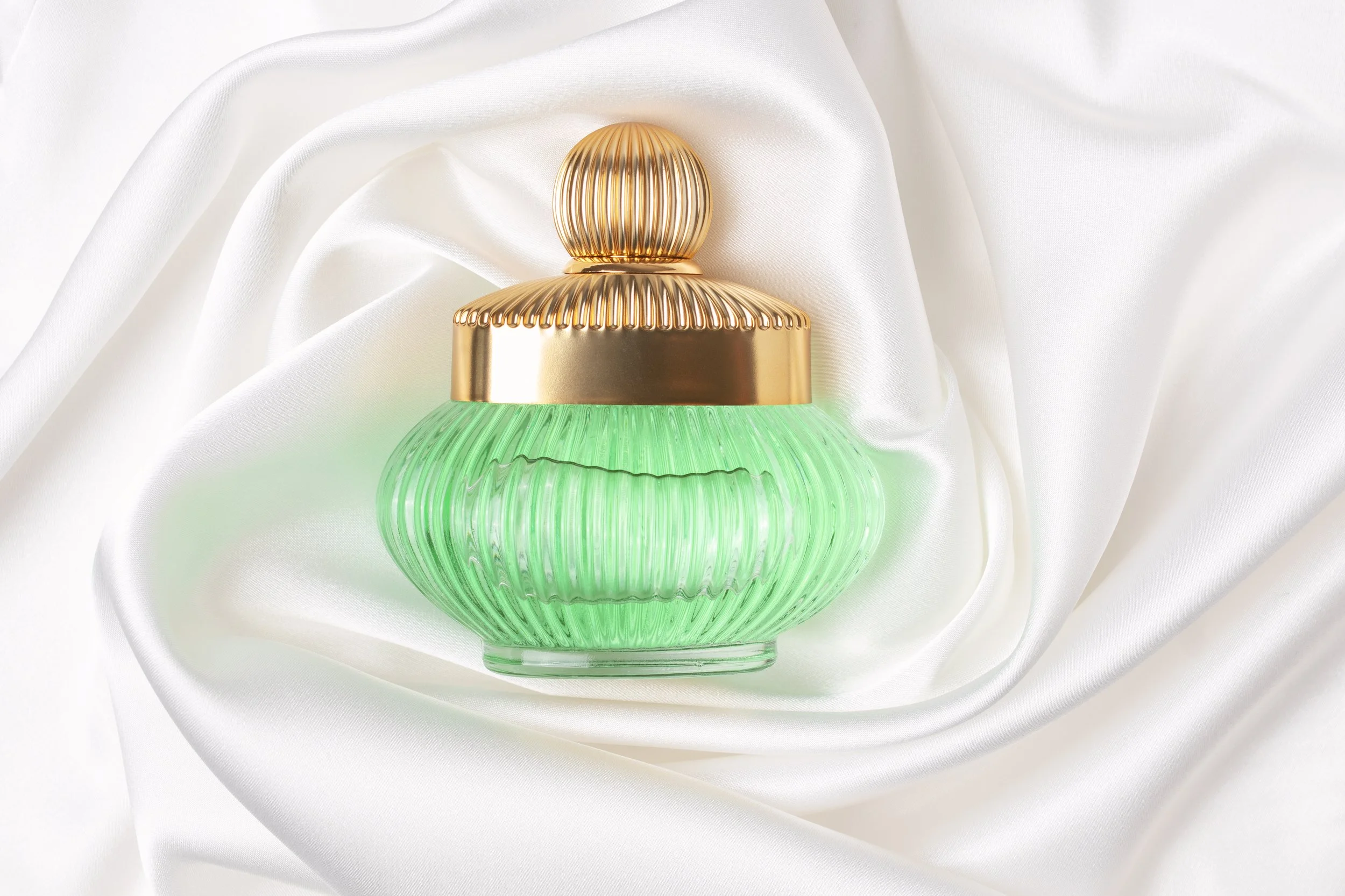 A decorative perfume bottle with a gold cap and a ribbed glass body in green, resting on white satin fabric.