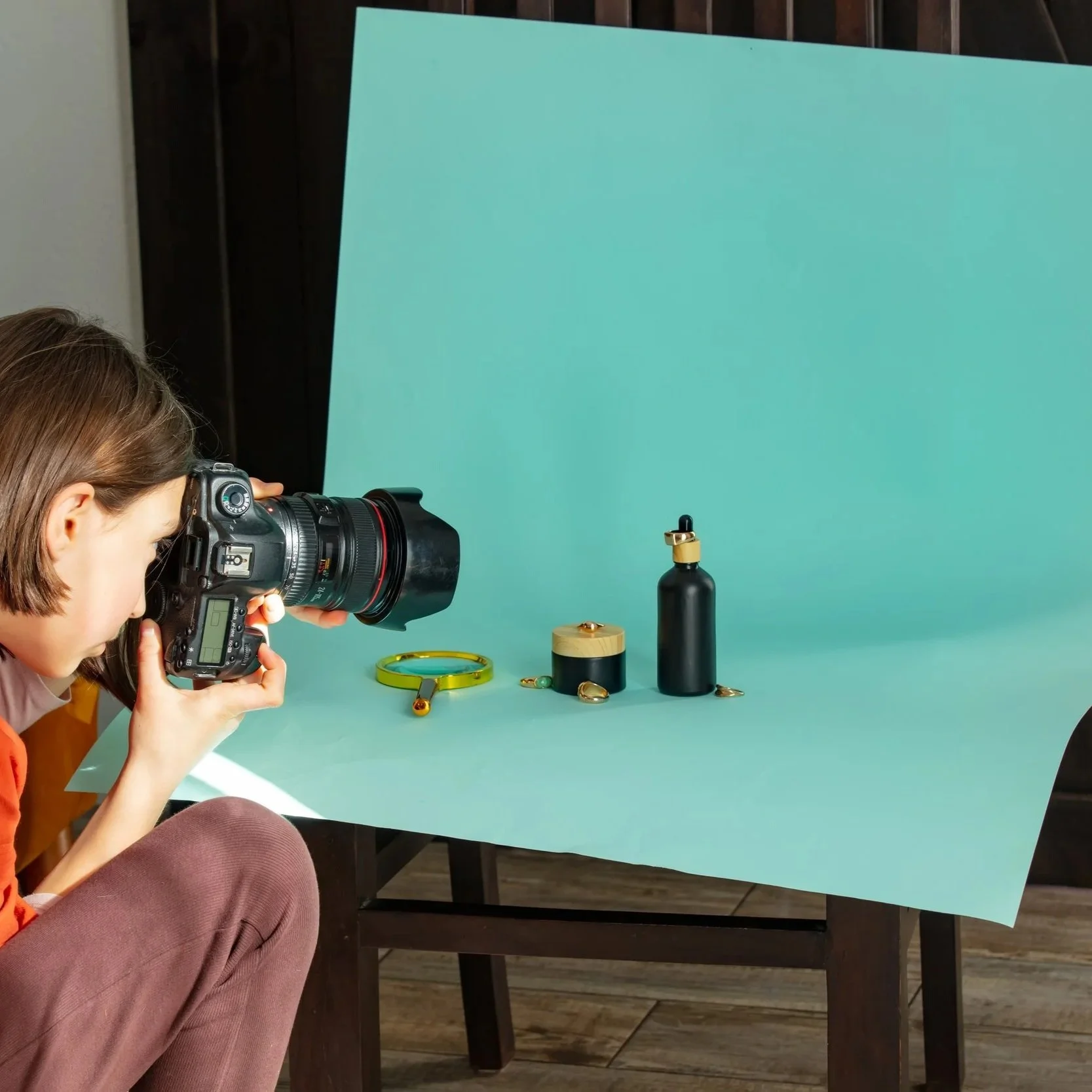 A person with shoulder-length brown hair taking a photograph of jewelry displayed on a tabletop with a light blue background.