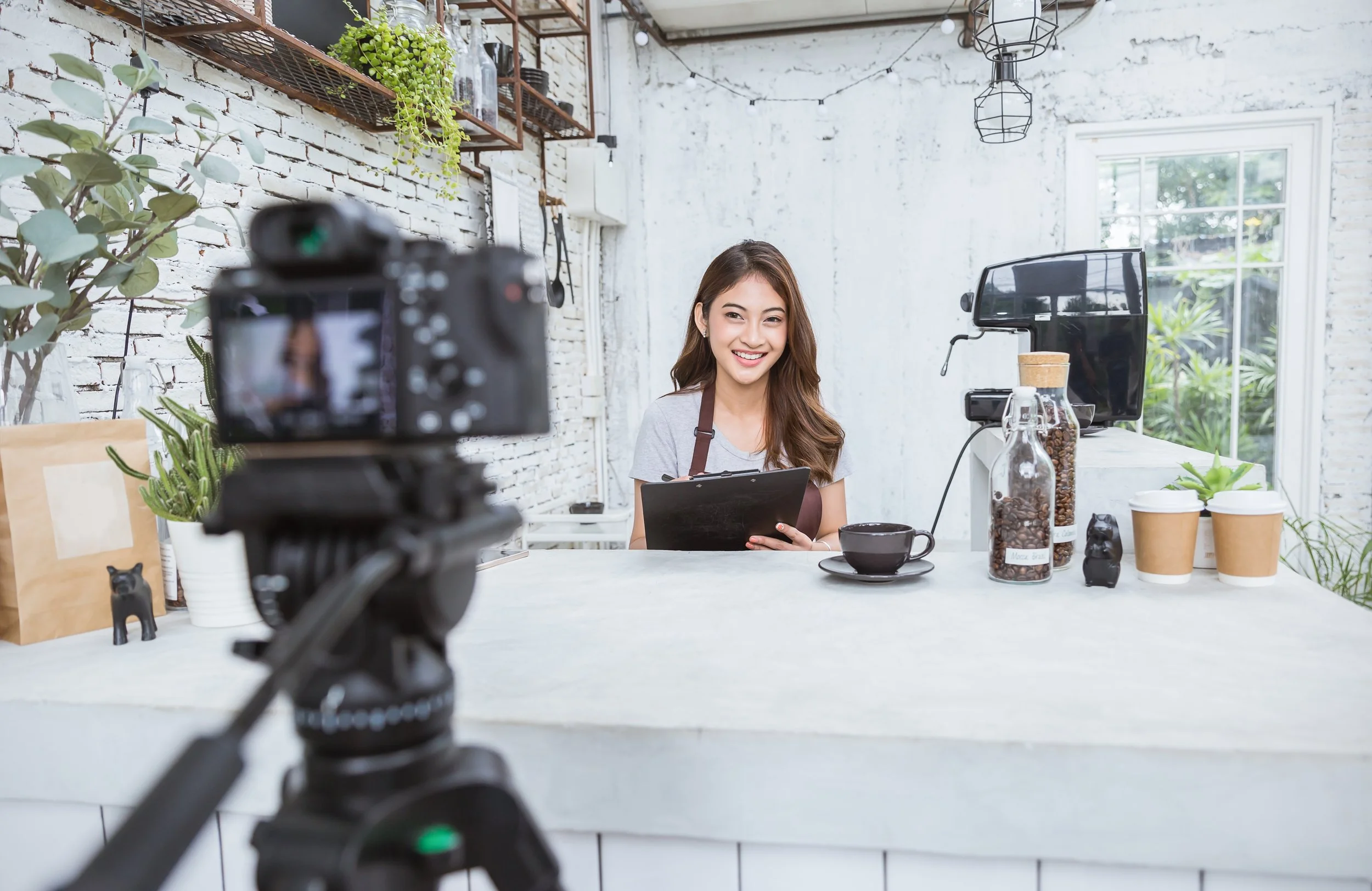 Young woman smiling at a cafe counter during a video recording, with camera focusing on her, surrounded by coffee beans, coffee cups, and potted plants in a bright, modern space with white brick walls and large windows.