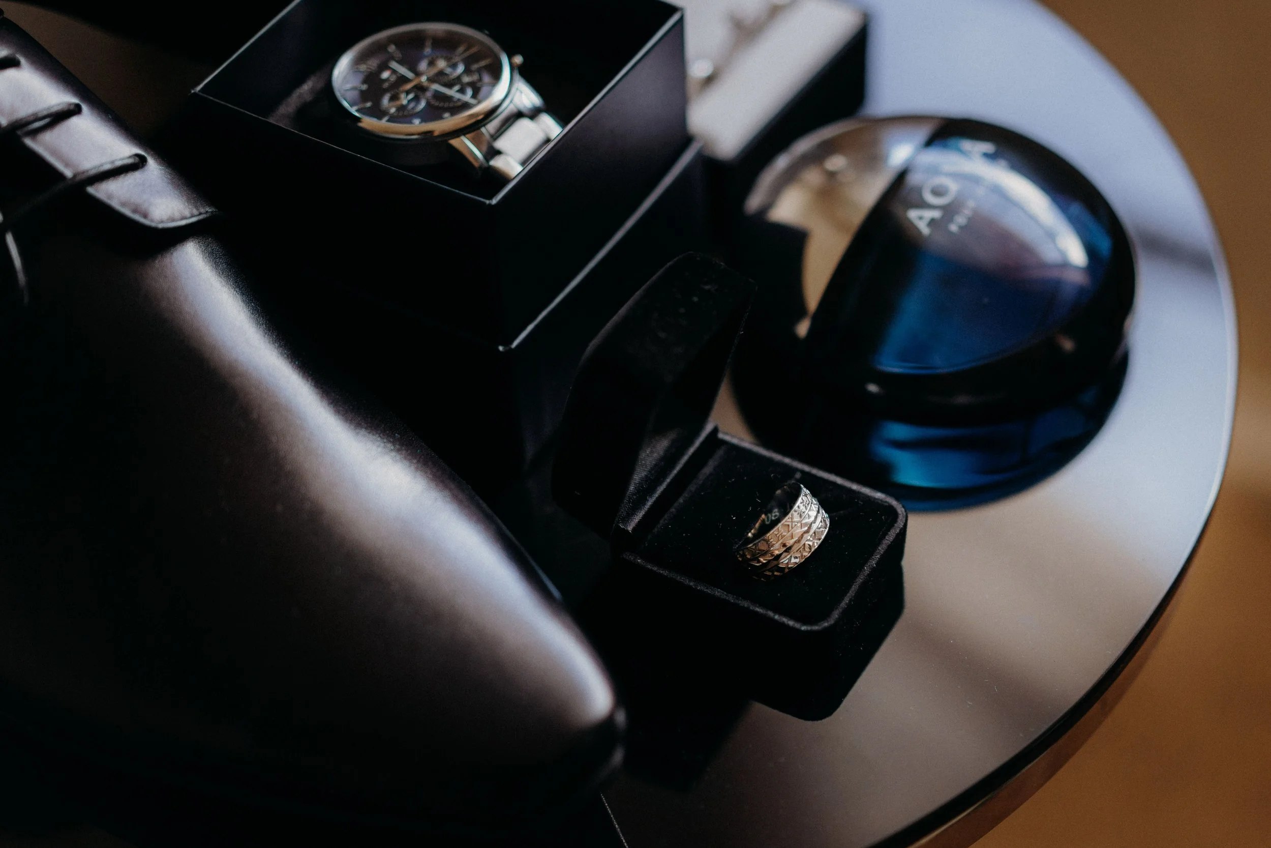 A close-up of a wristwatch in a box, a ring in a velvet holder, and a container of AOH perfume on a reflective surface.