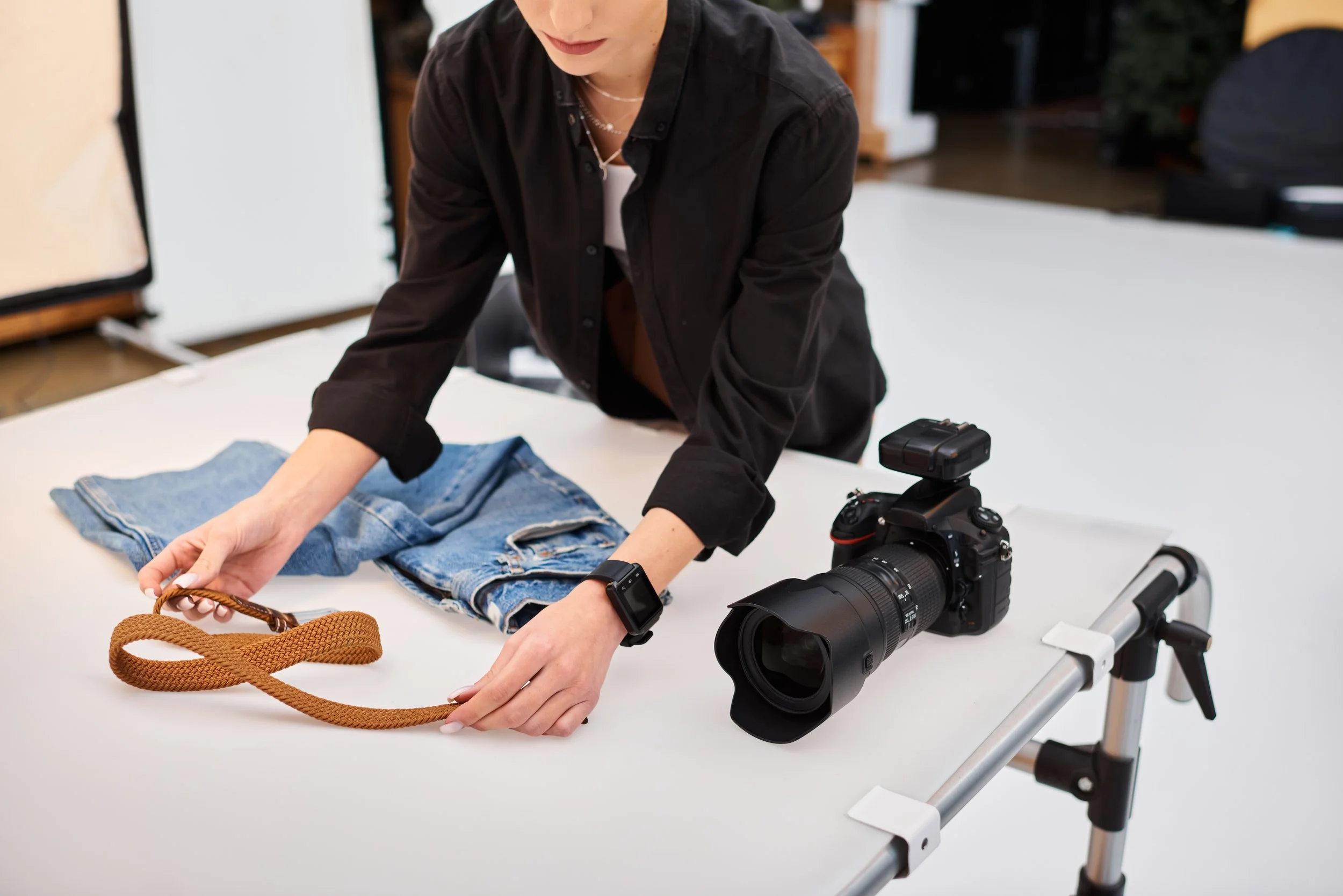 Person arranging a leather belt, a pair of denim jeans, a professional camera with a large lens on a white table in a studio.