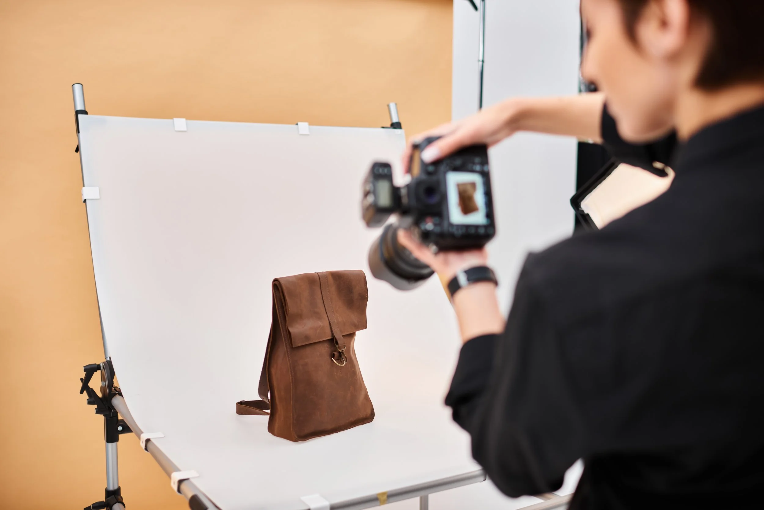 A person taking a photo of a brown leather bag on a white photography backdrop with a camera.