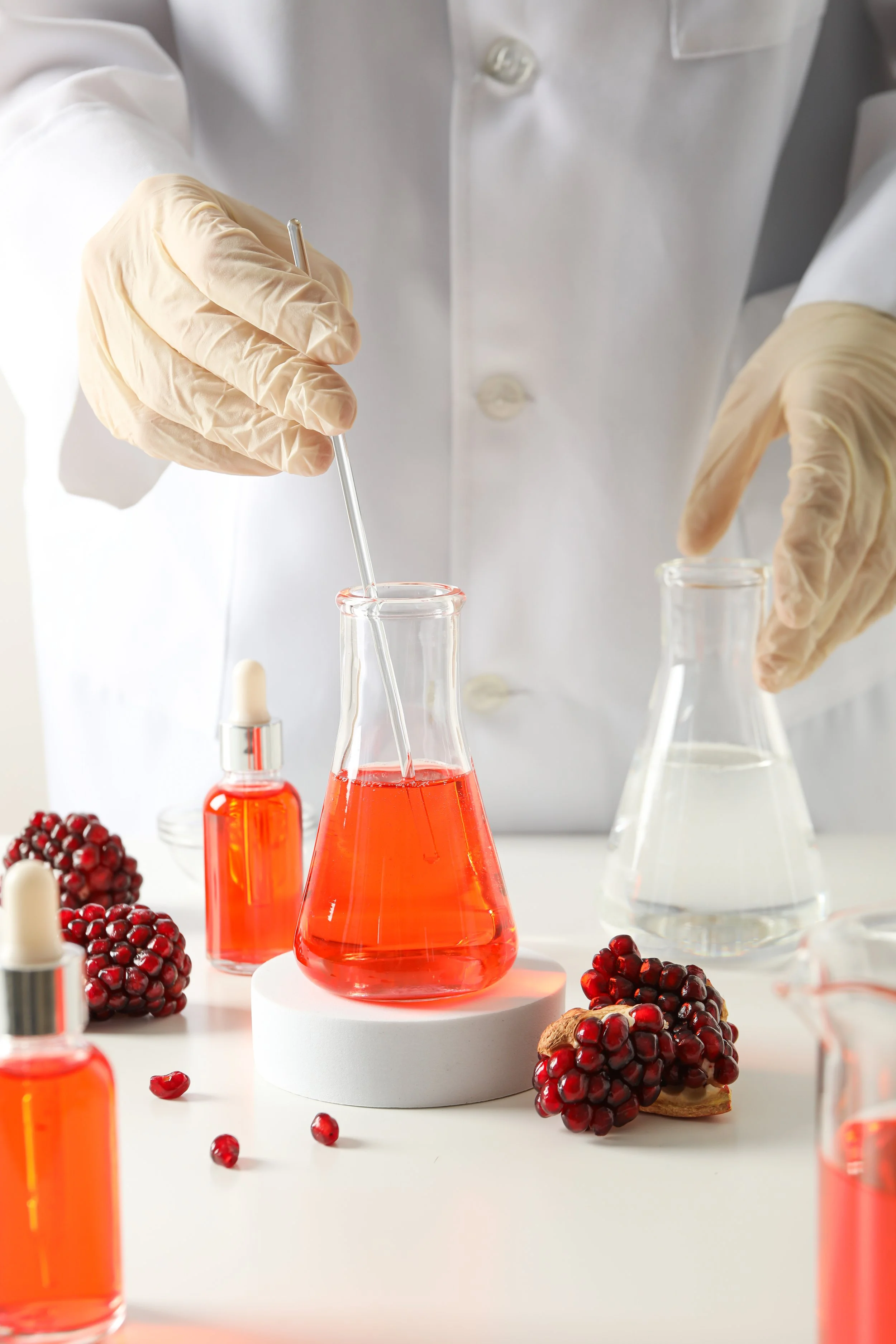 Person in a lab coat using a glass pipette to transfer red liquid into a glass test tube, surrounded by laboratory glassware and pomegranate fruit.