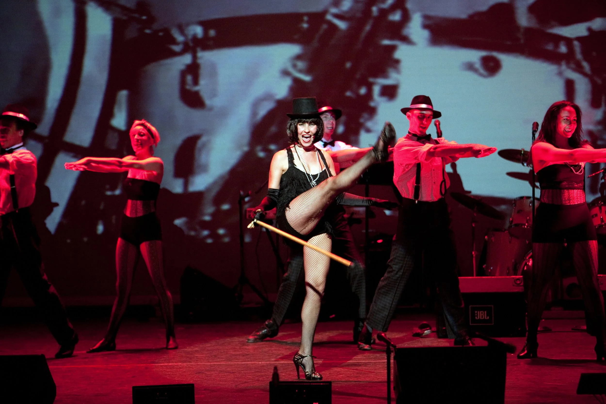 Performer in a black costume with fishnet tights, a black top hat, and holding a cane, dancing with one leg high in the air surrounded by backup dancers dressed in red suspenders and black hats, on a stage with a large black and white photo projected
