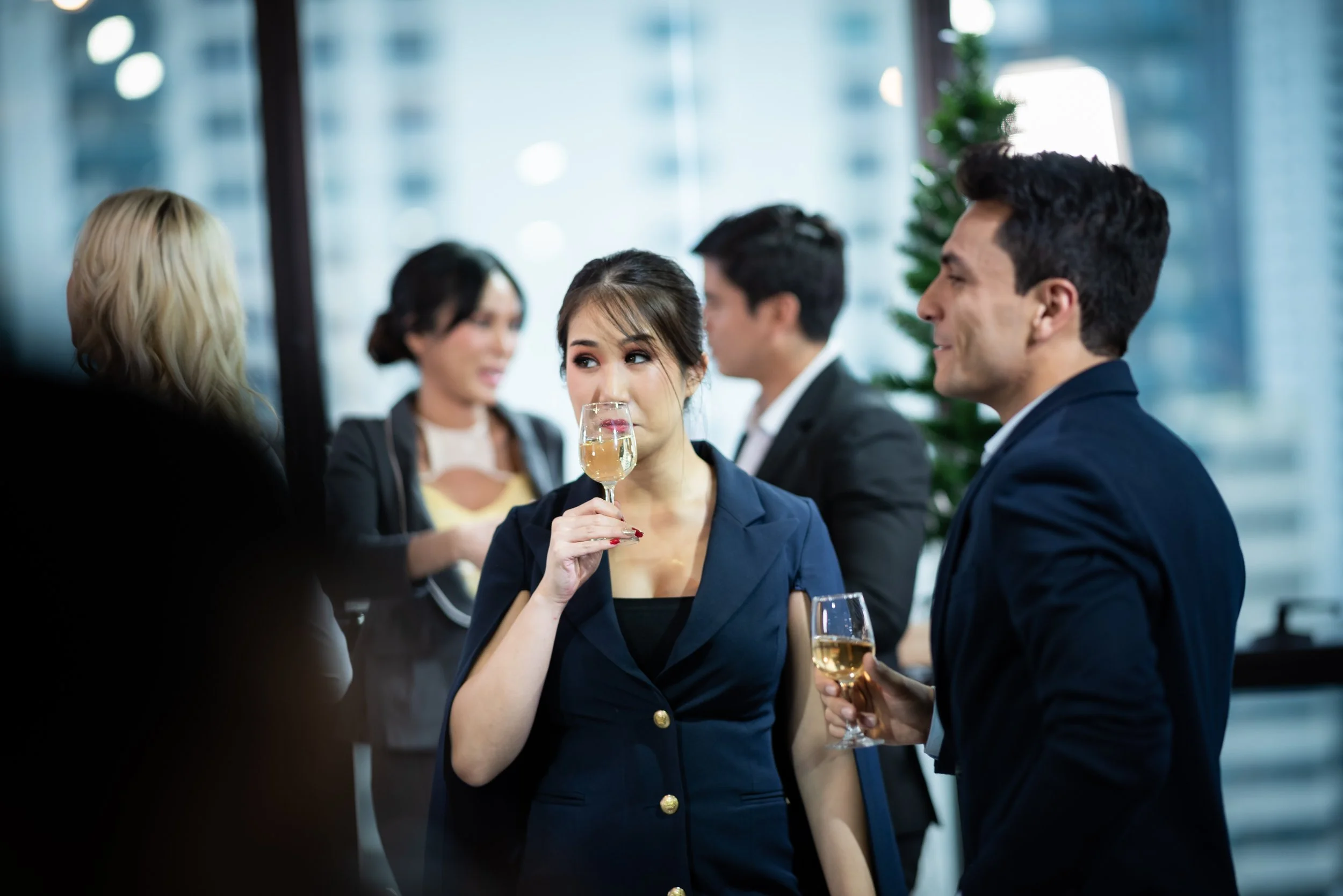 Business professionals at a social gathering, with a woman sipping champagne and a man holding a glass of wine, in an office setting with large windows and cityscape in the background.