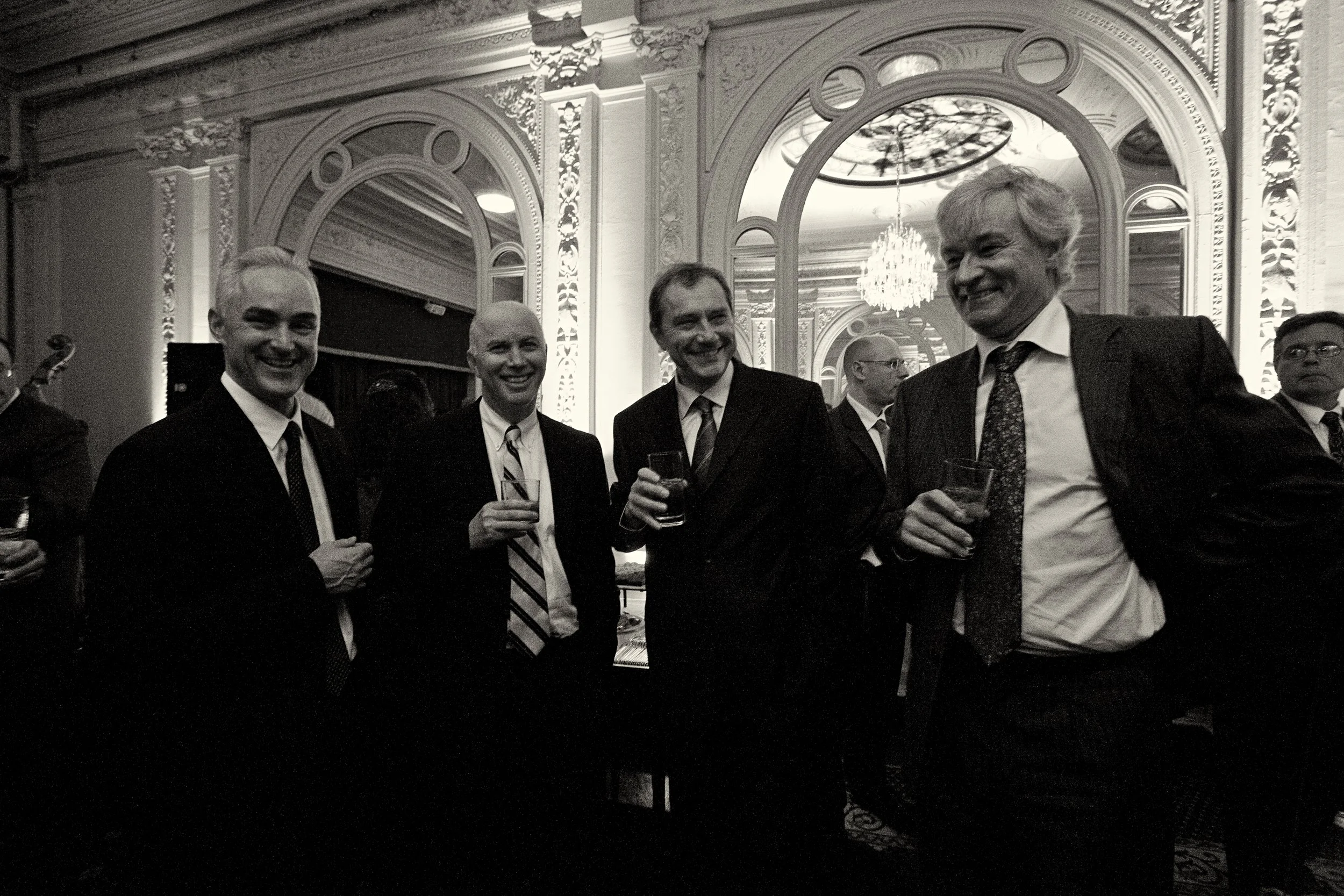 A group of five men in suits at a formal event, holding drinks and smiling, with an ornate room featuring large mirrors and chandeliers.