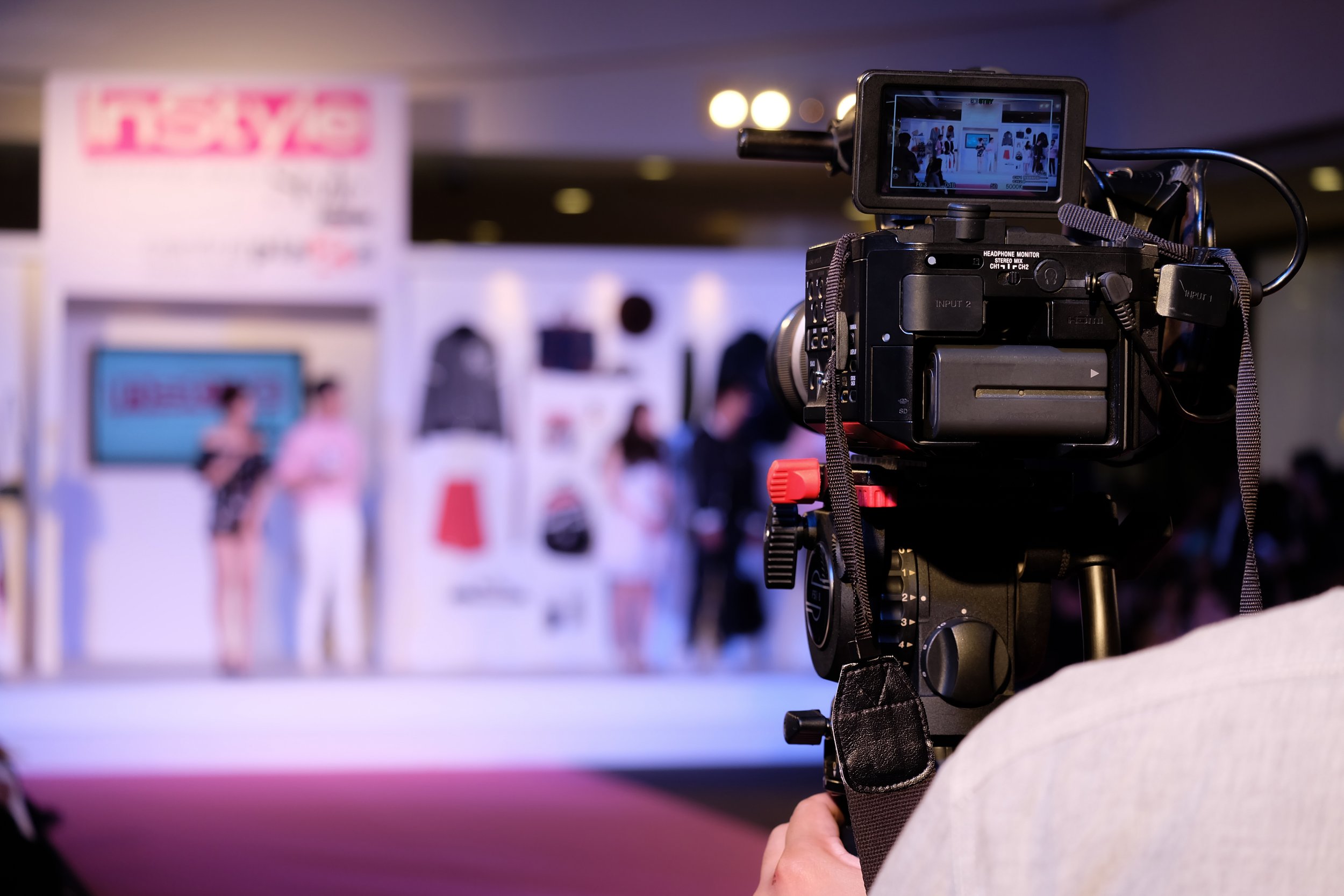 A professional video camera recording a fashion show or presentation with models on stage, in a well-lit indoor venue.