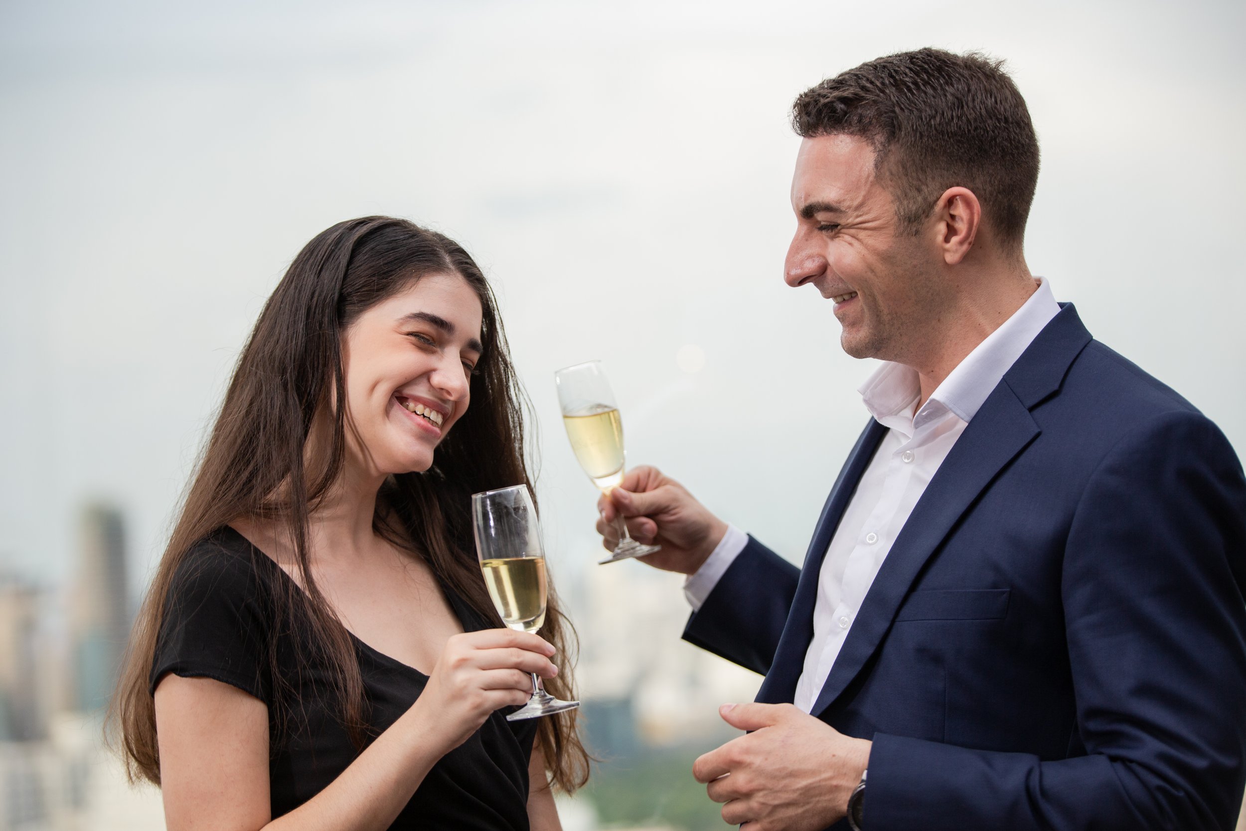 A man and woman celebrating with champagne glasses, both smiling and dressed in formal attire, against a city skyline background.