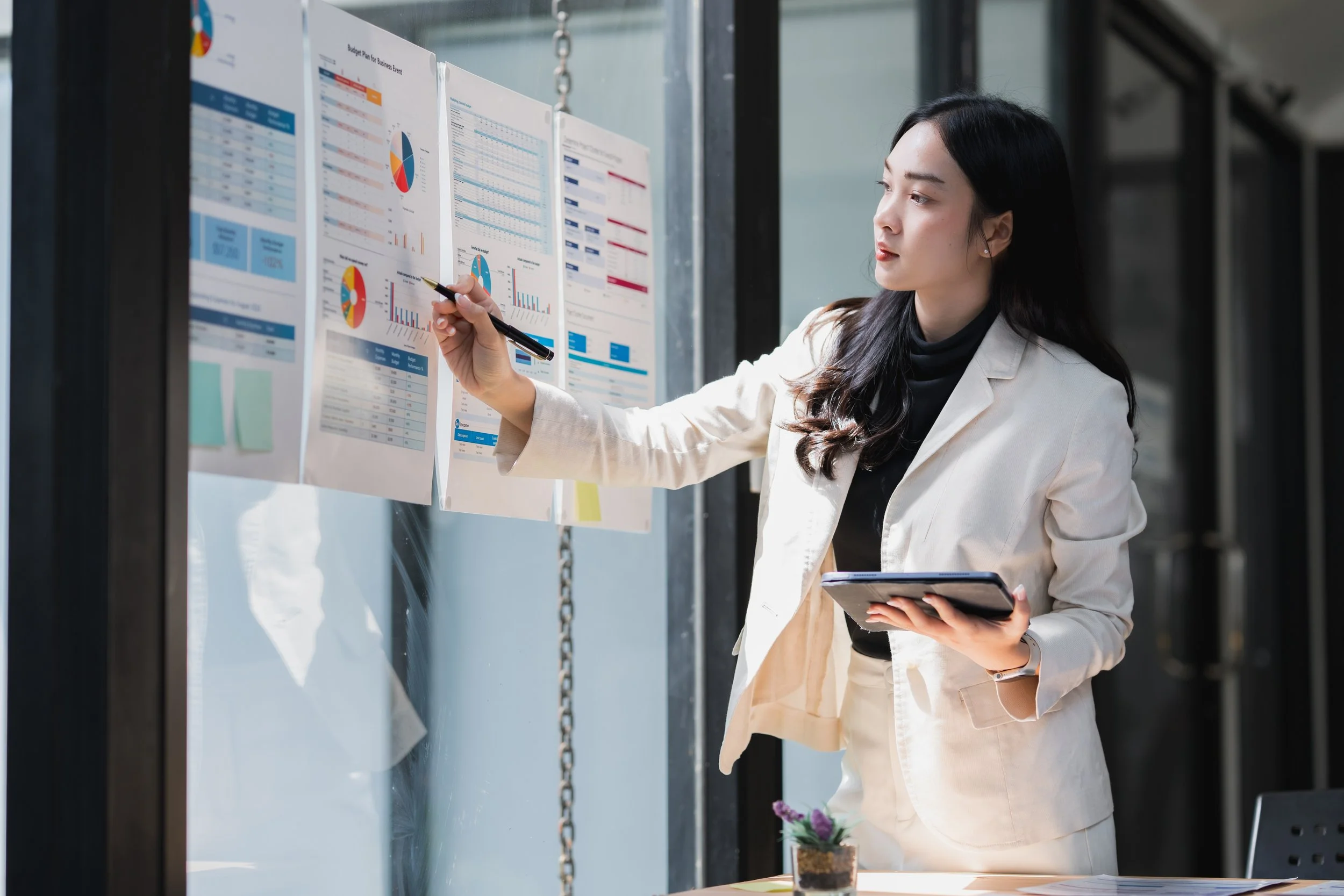 A woman in a white blazer and black turtleneck pointing at papers with charts and graphs on a glass wall, holding a tablet in her left hand.
