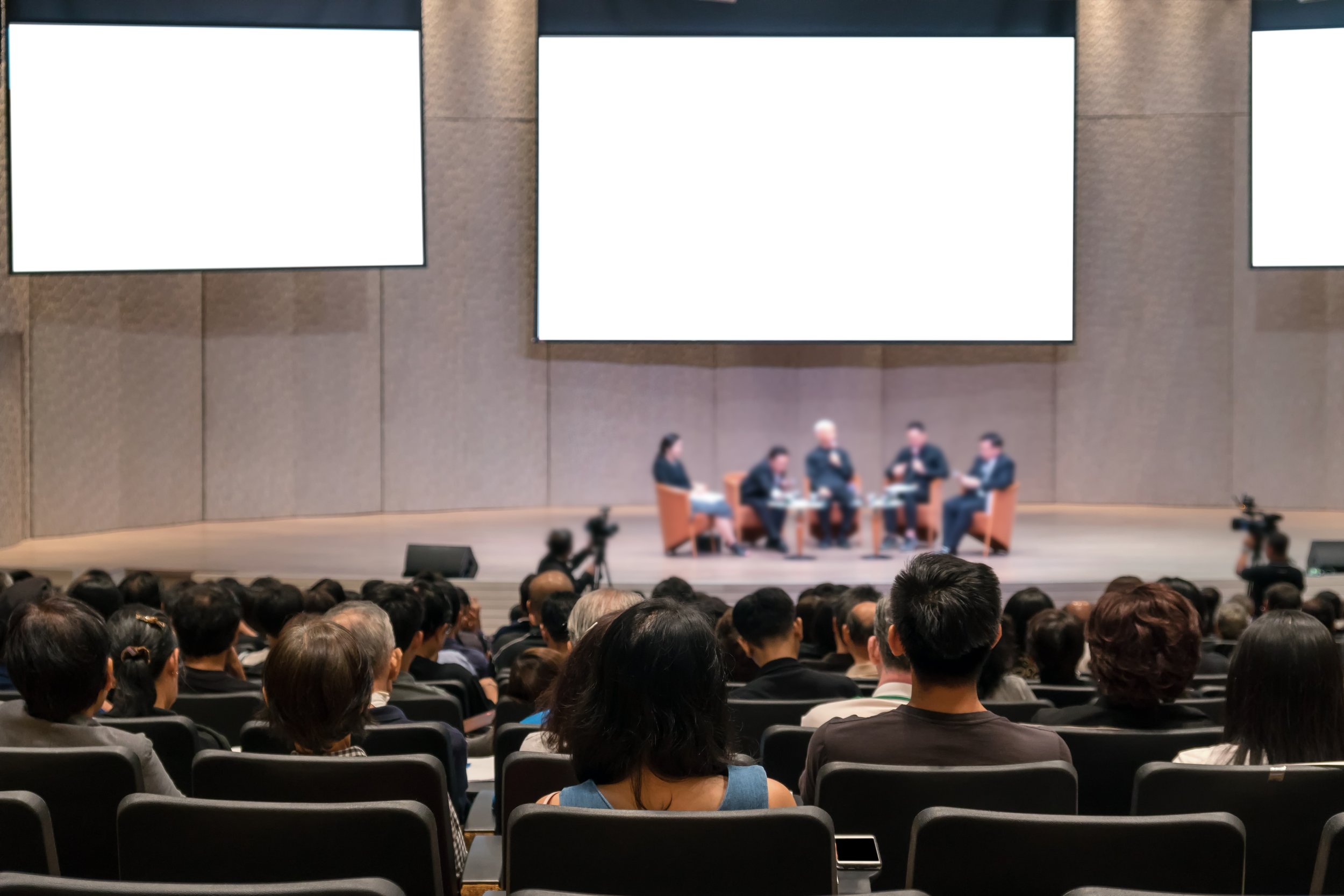 An audience watching a panel discussion on stage with five speakers in a conference hall.