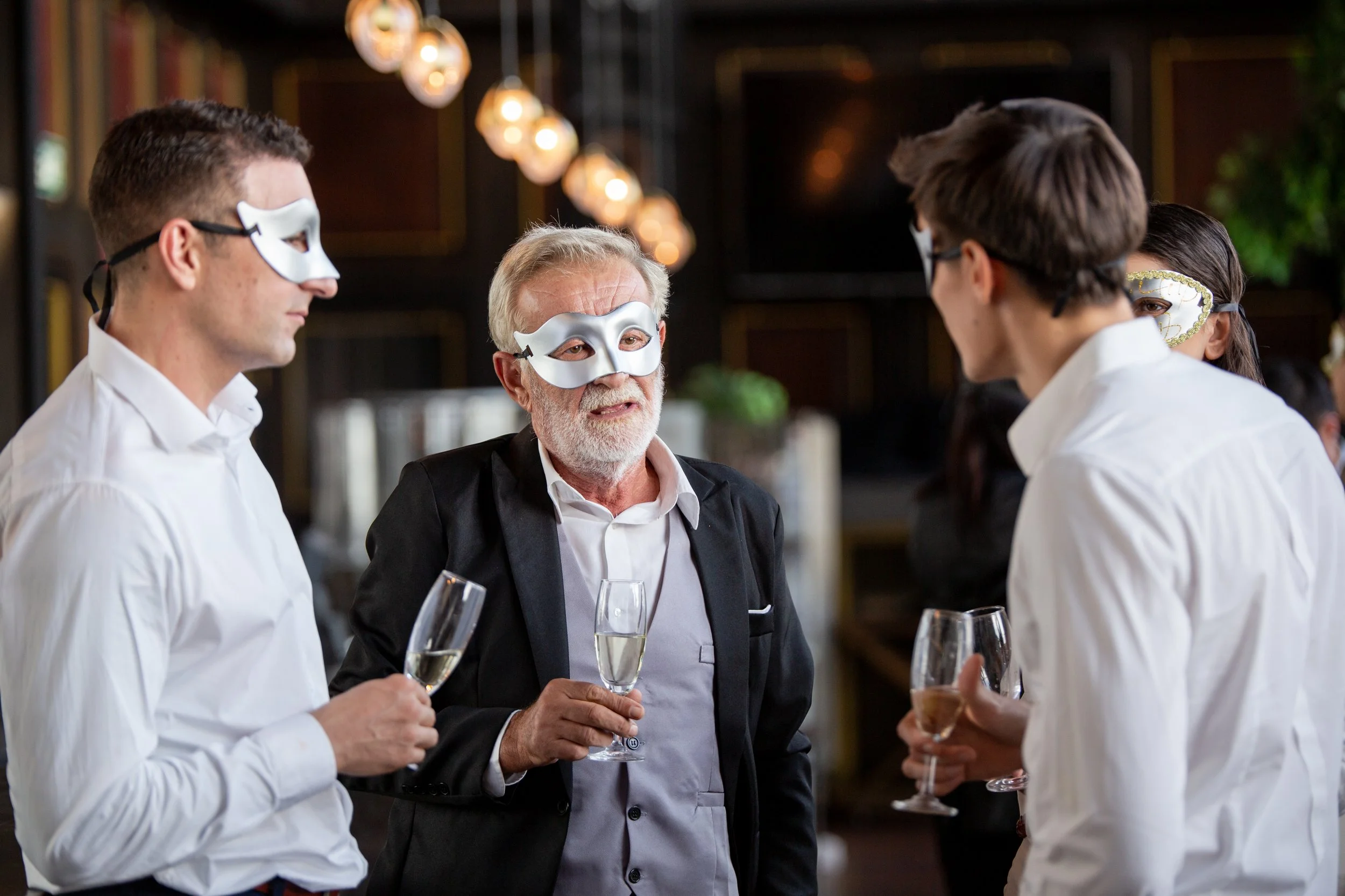 A group of people wearing masquerade masks and formal white shirts, holding glasses of champagne, socializing at a party.