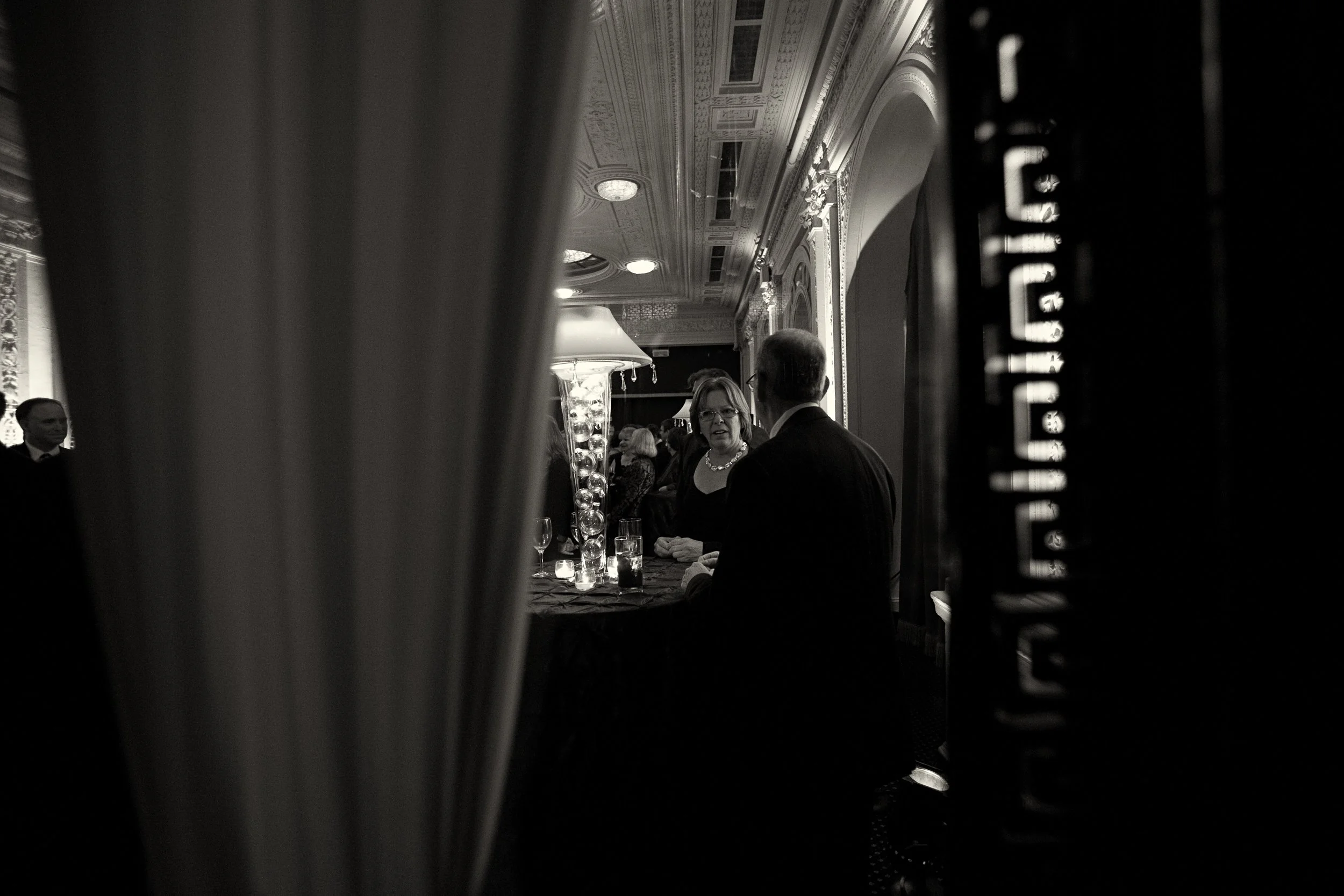 A black and white photo captures a man and a woman engaged in conversation at a formal event in an ornate room with decorative ceiling moldings and draped curtains.