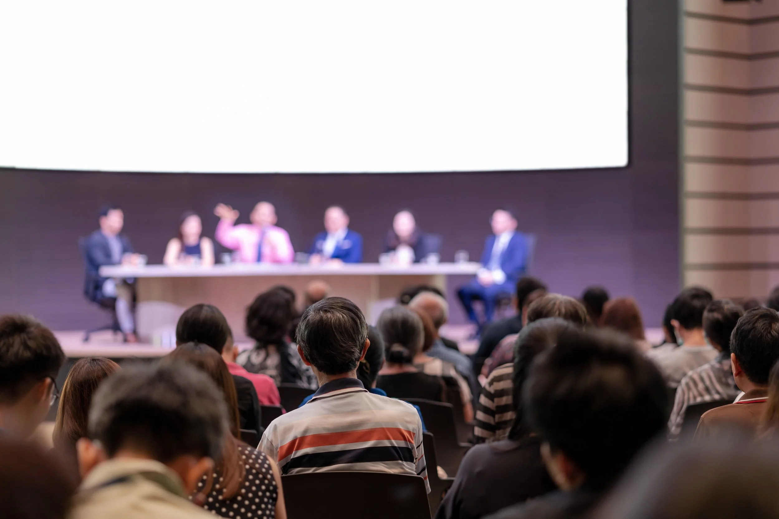 A panel of six people sitting at a long table on a stage, with one person talking and gesturing. An audience is seated facing them in a conference room.