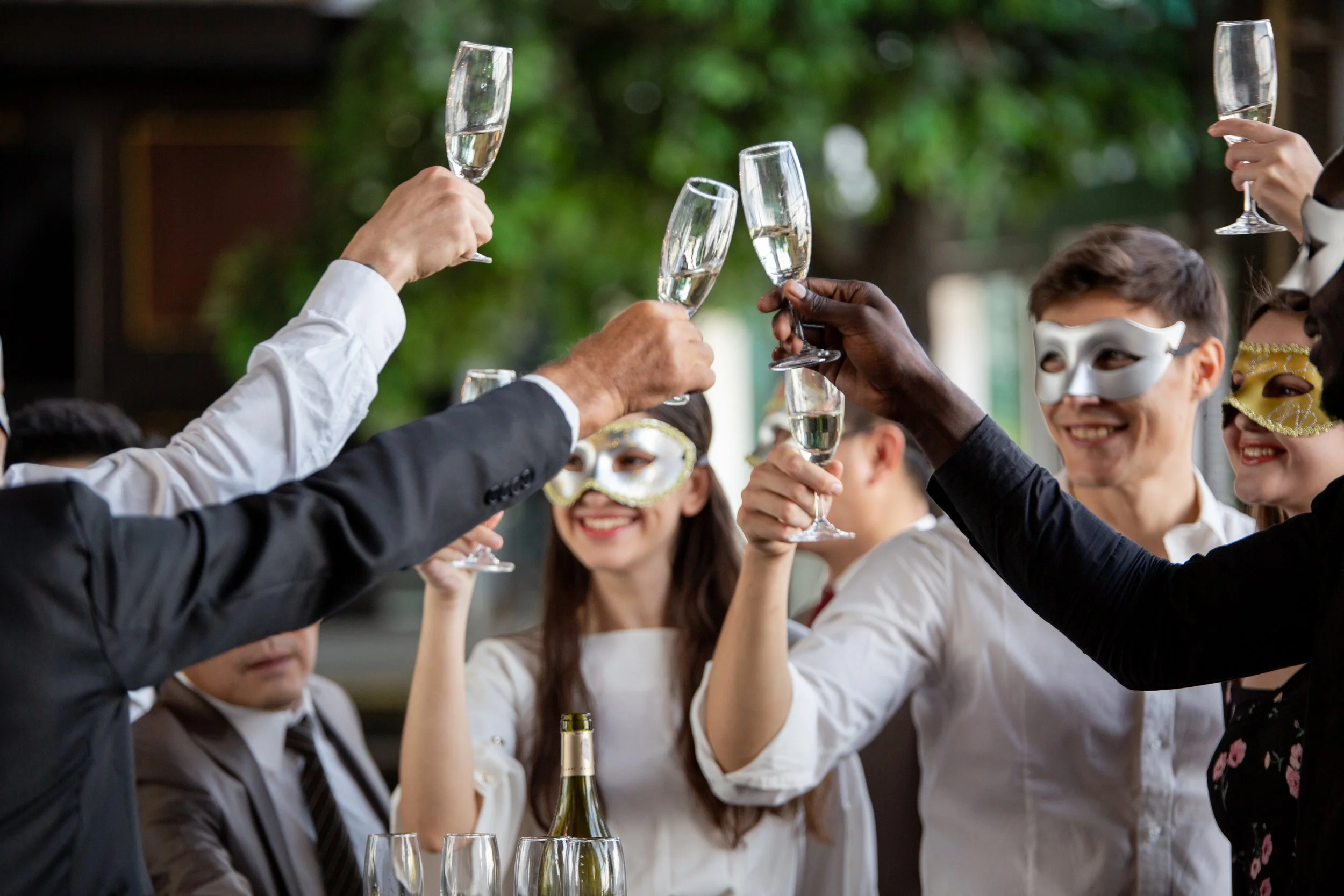 People celebrating with glasses of champagne, some wearing masquerade masks, during a festive outdoor gathering.