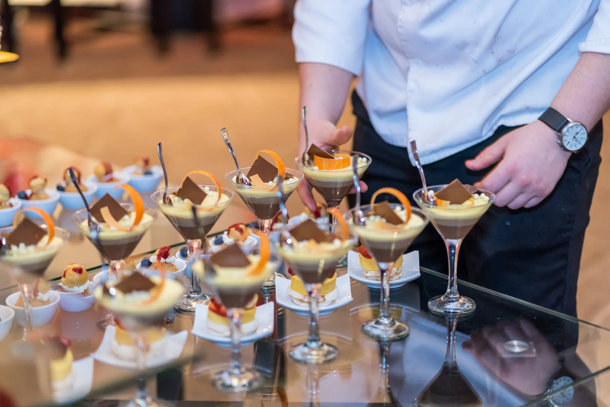 A person in a white shirt and black pants preparing a tray of assorted desserts, including chocolate mousse in martini glasses and small pastries, on a glass table.