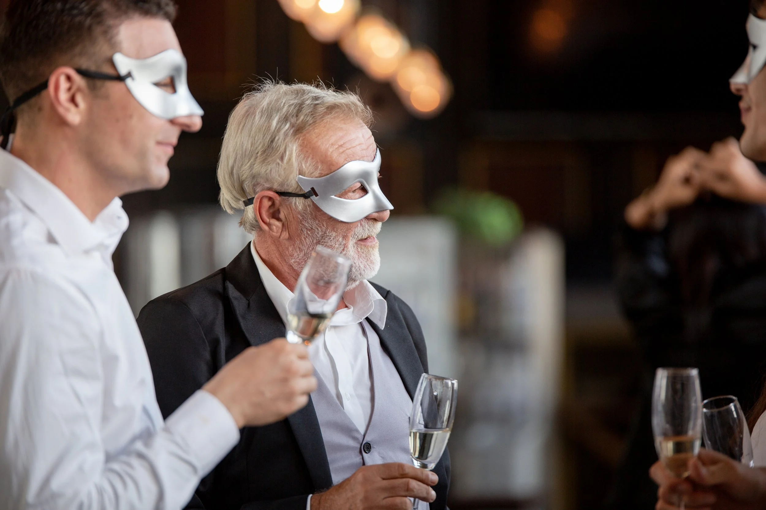 People at a party wearing silver masks, holding champagne glasses, and engaging in conversation in a dimly lit setting.
