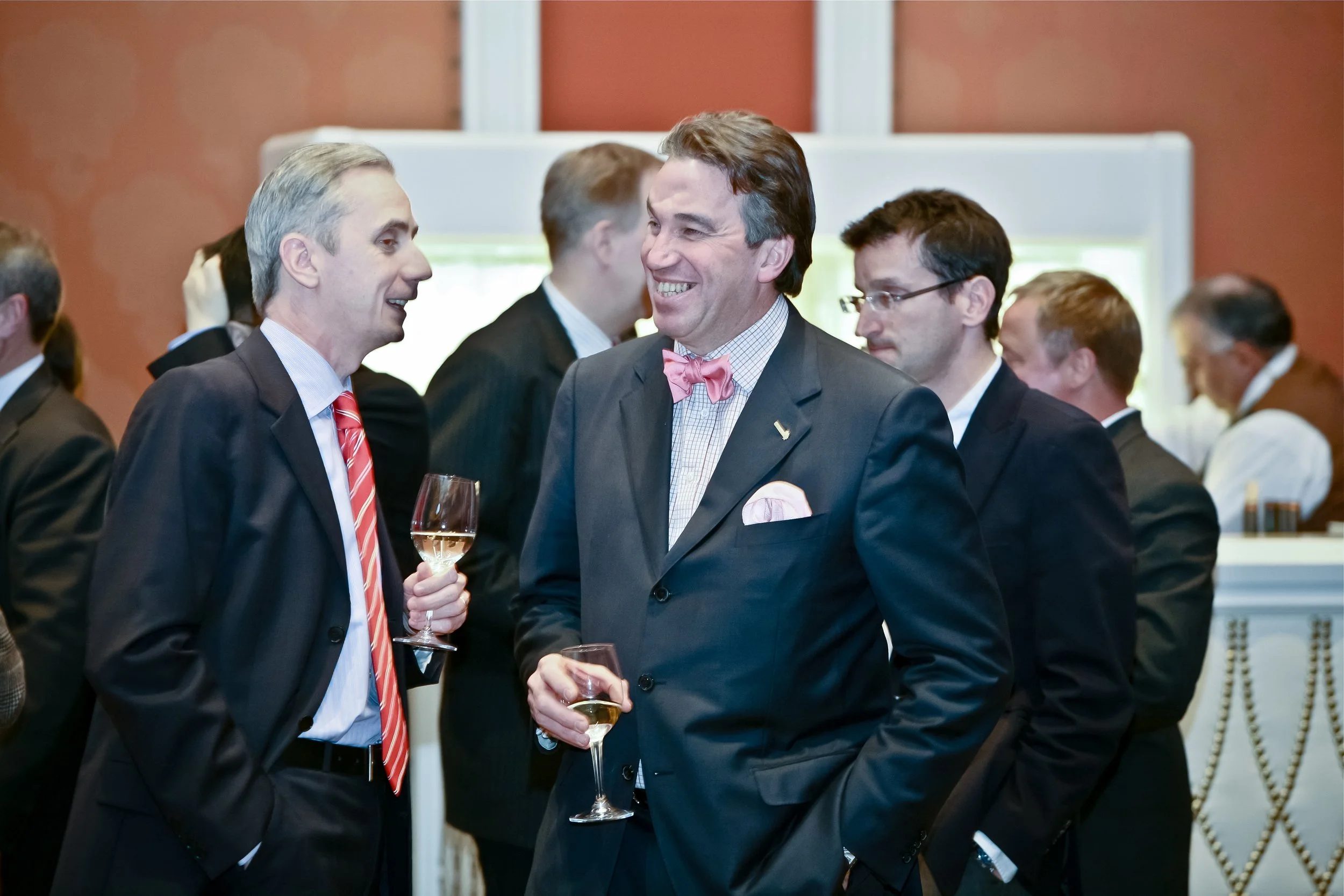 Two men in suits and pink accessories engaged in conversation at a formal event, holding wine glasses, with other attendees in the background.