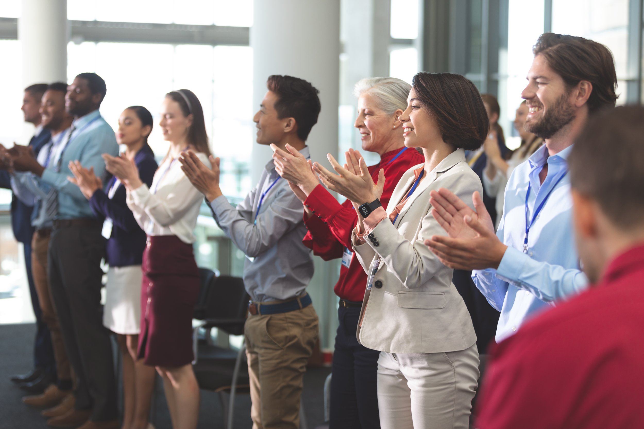 A diverse group of people attending a conference or seminar, clapping and smiling in a modern, well-lit venue with large windows.