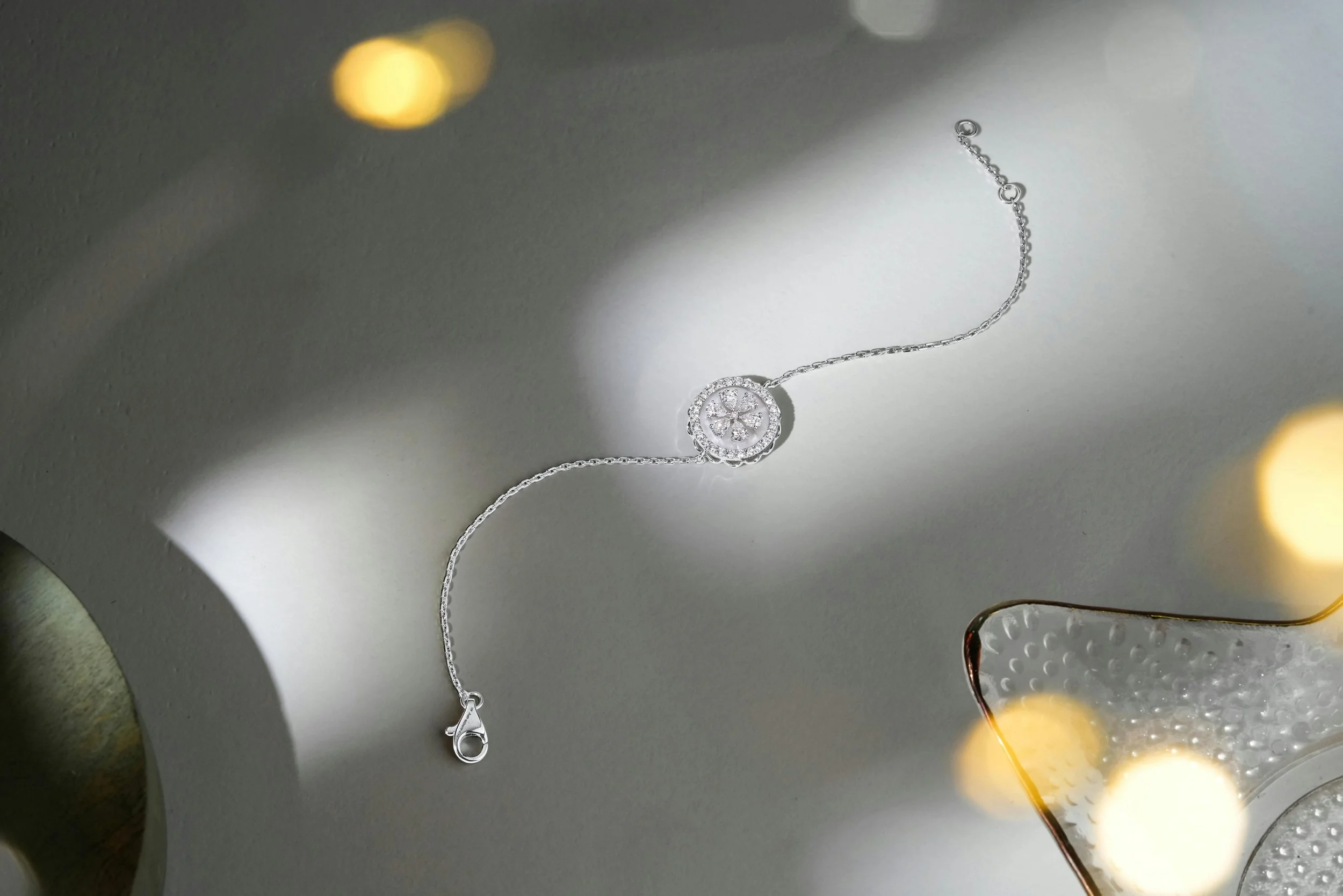 A silver necklace with a circular, jeweled pendant resting on a gray surface, with a glass dish and blurred warm lights in the background.