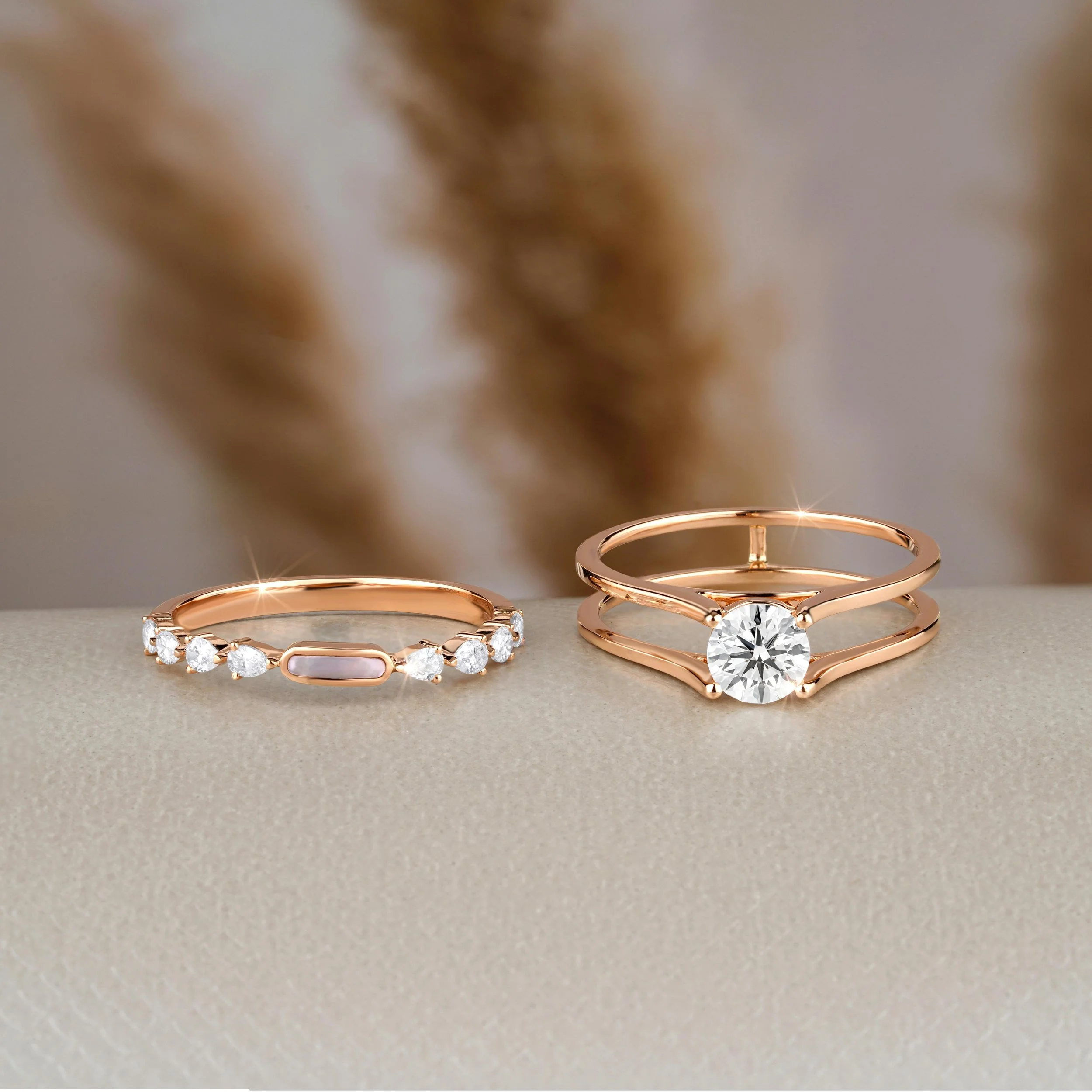 Two rose gold rings, one with a large central diamond and a thin band, and the other with a row of small diamonds and a rectangular pink gemstone, on a neutral surface with a blurred background.