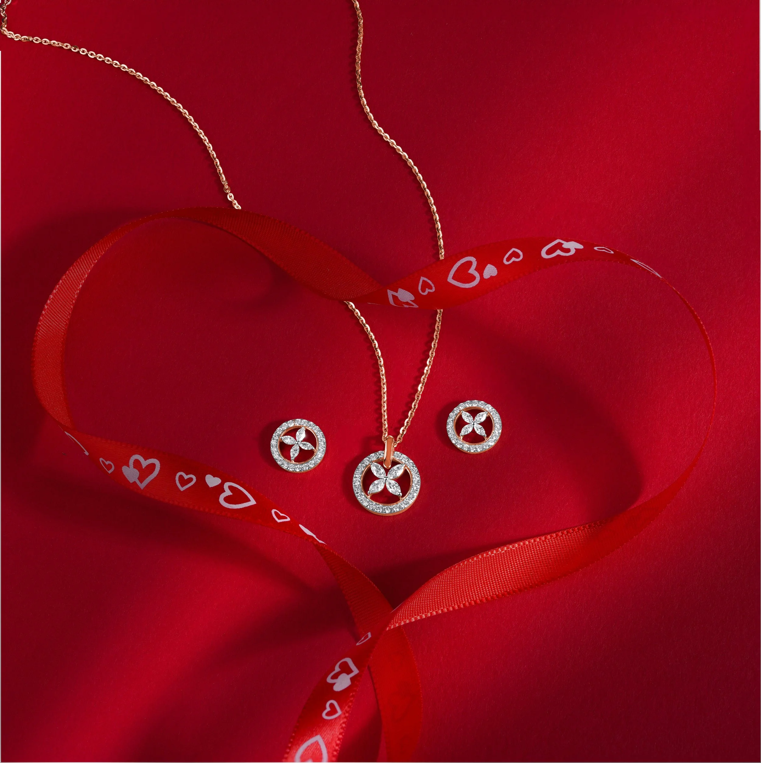 Gold necklace and earrings with circular pendants decorated with small diamonds, displayed on red background with a red ribbon featuring white hearts and card suit symbols, arranged to form a heart shape.