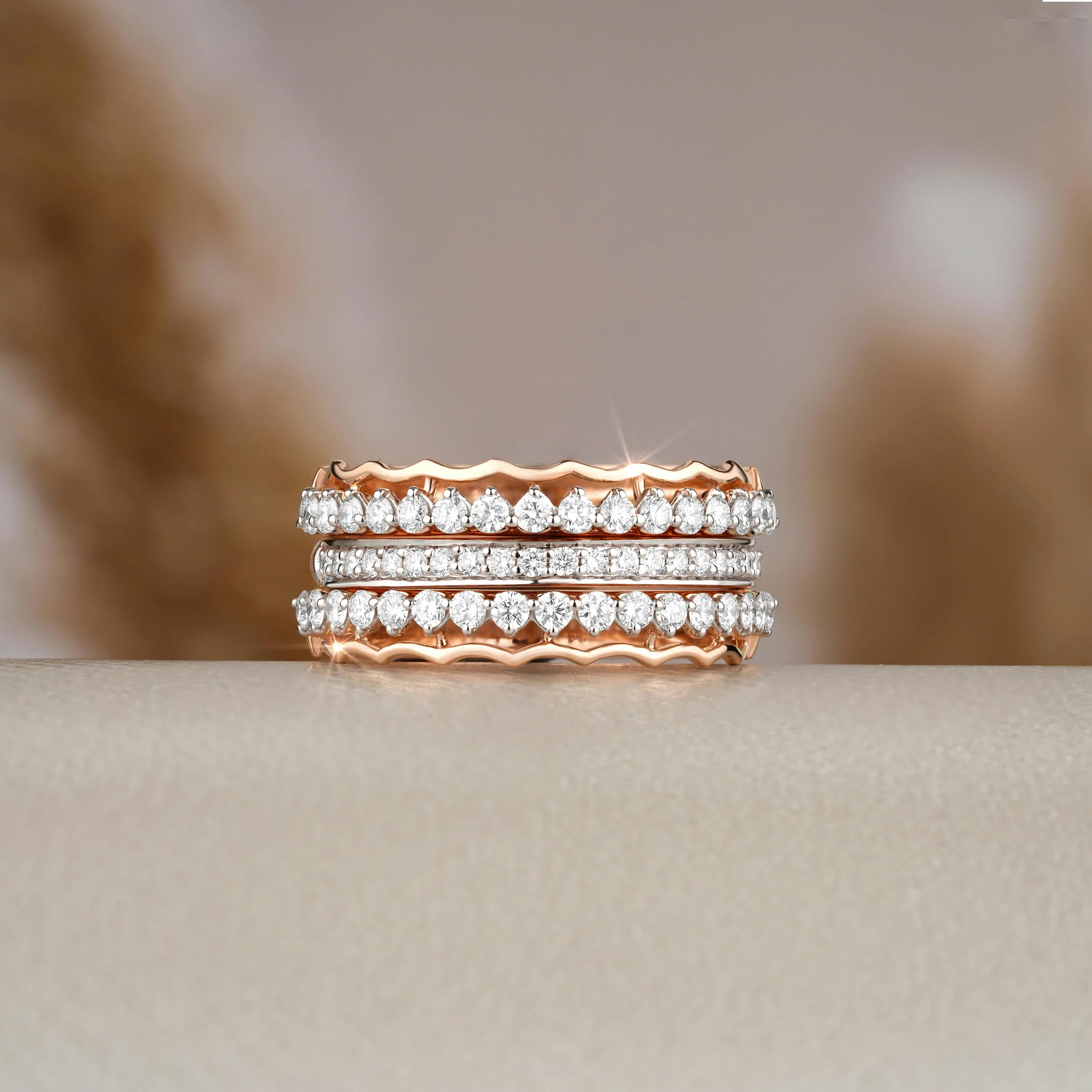 A stack of three rings with embedded diamonds, with a wavy design, placed on a beige surface with a blurred background.