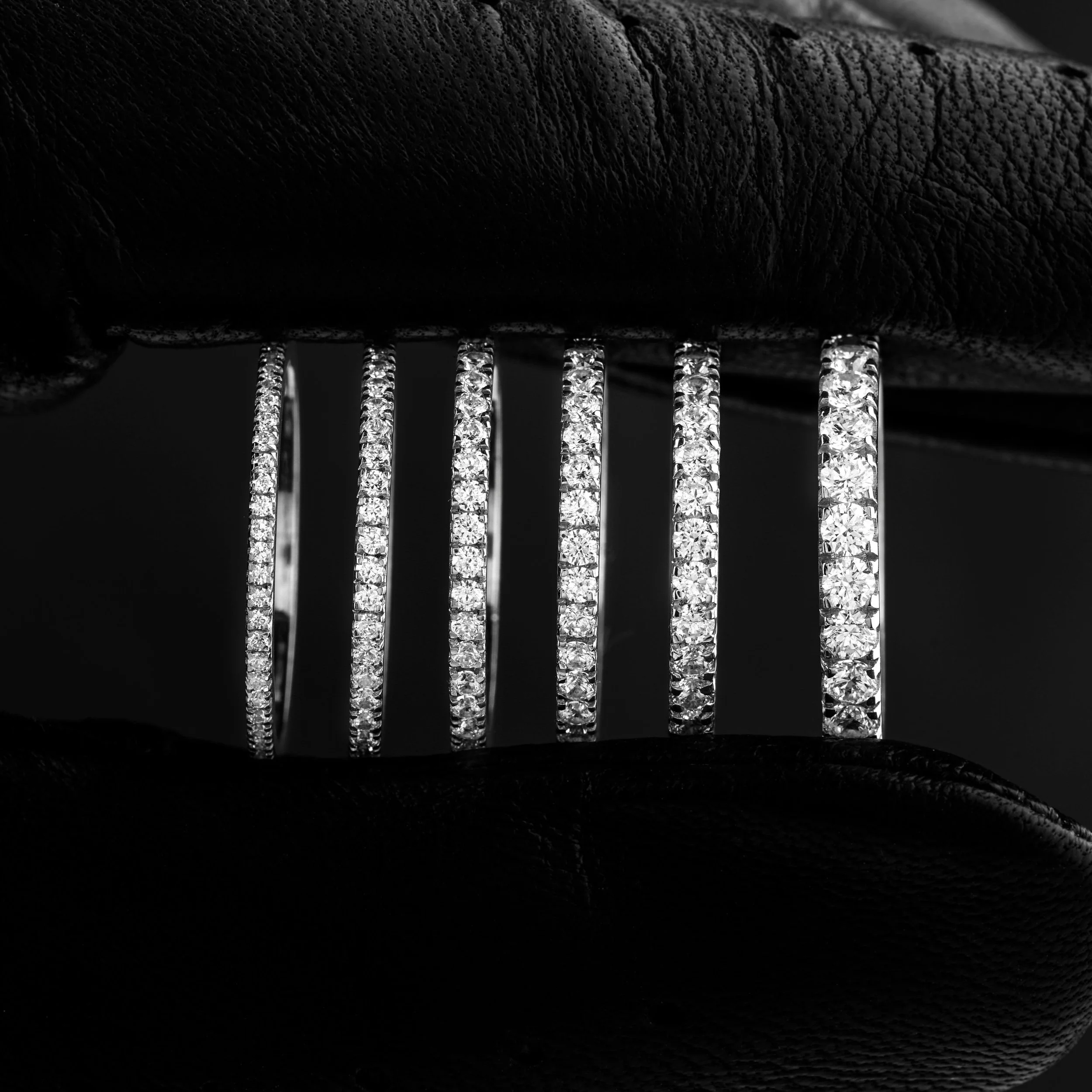 Seven diamond-studded rings held between fingers wearing black leather gloves, reflected on a glossy black surface.