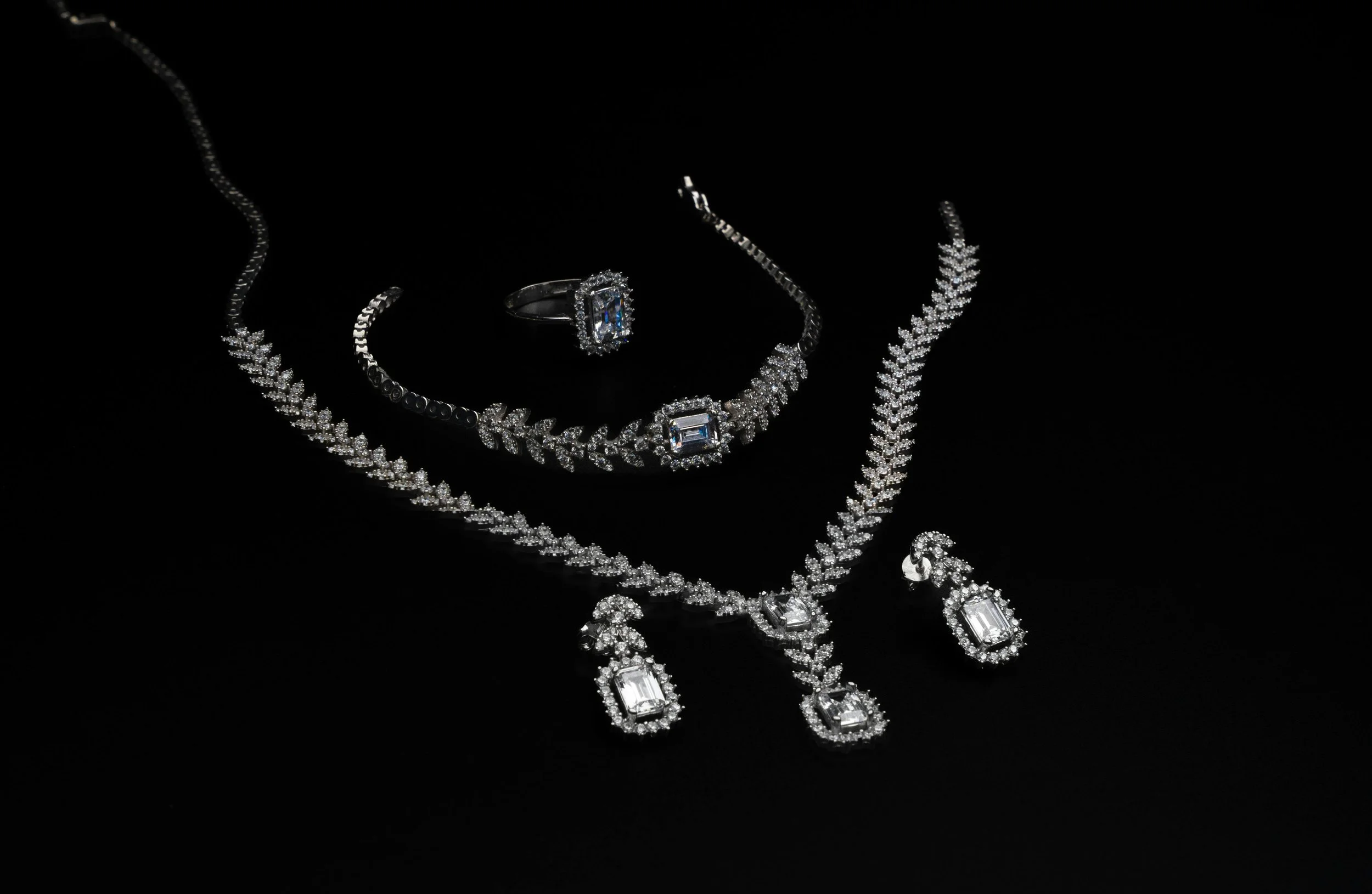 Elegant jewelry set on black background, including a necklace, bracelet, ring, and earrings, all adorned with sparkling diamonds in various cuts.