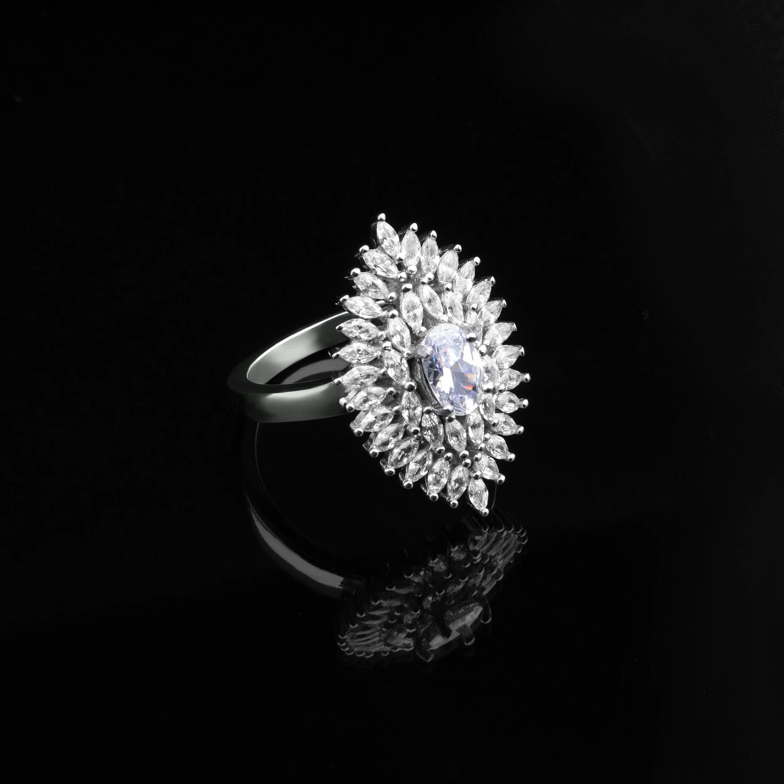 Diamond cocktail ring with a large central oval diamond surrounded by smaller marquise and round diamonds, set on a silver band, against a black background with reflection.