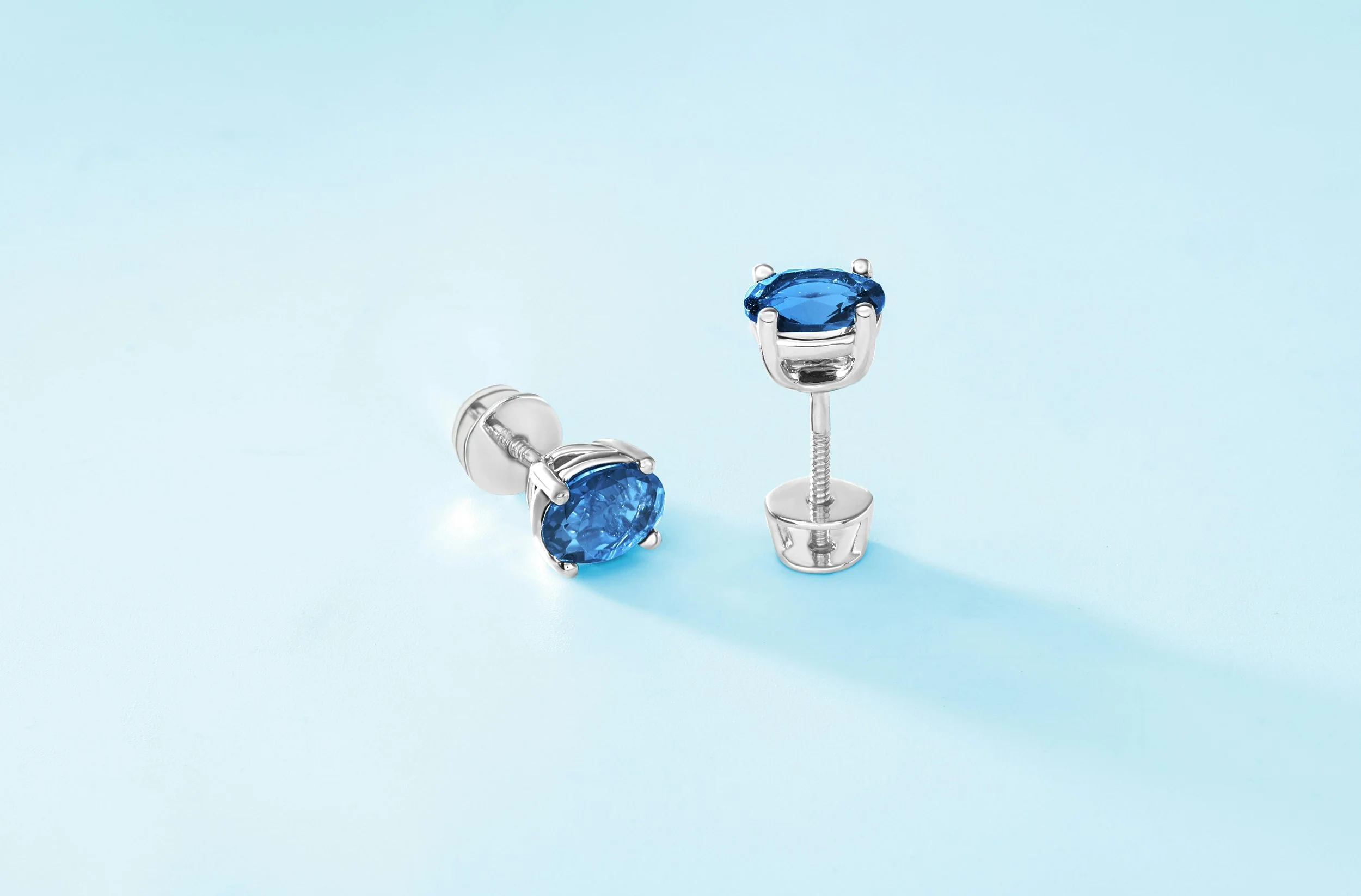 Pair of silver stud earrings with blue gemstones on a light blue background, one earring lying down and the other standing upright.