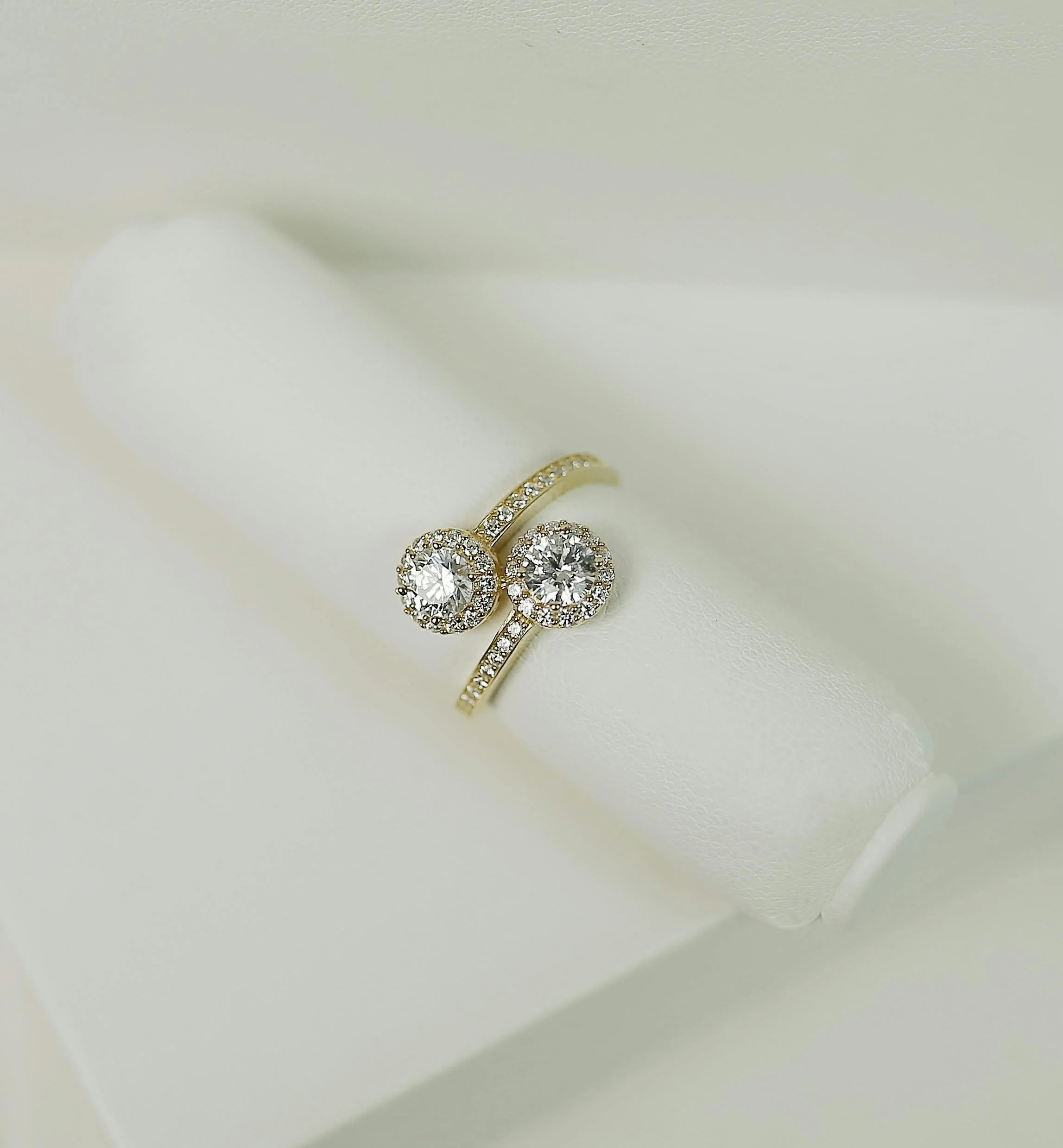 A gold ring with two round diamond center stones surrounded by smaller diamonds, displayed on a white jewelry box.