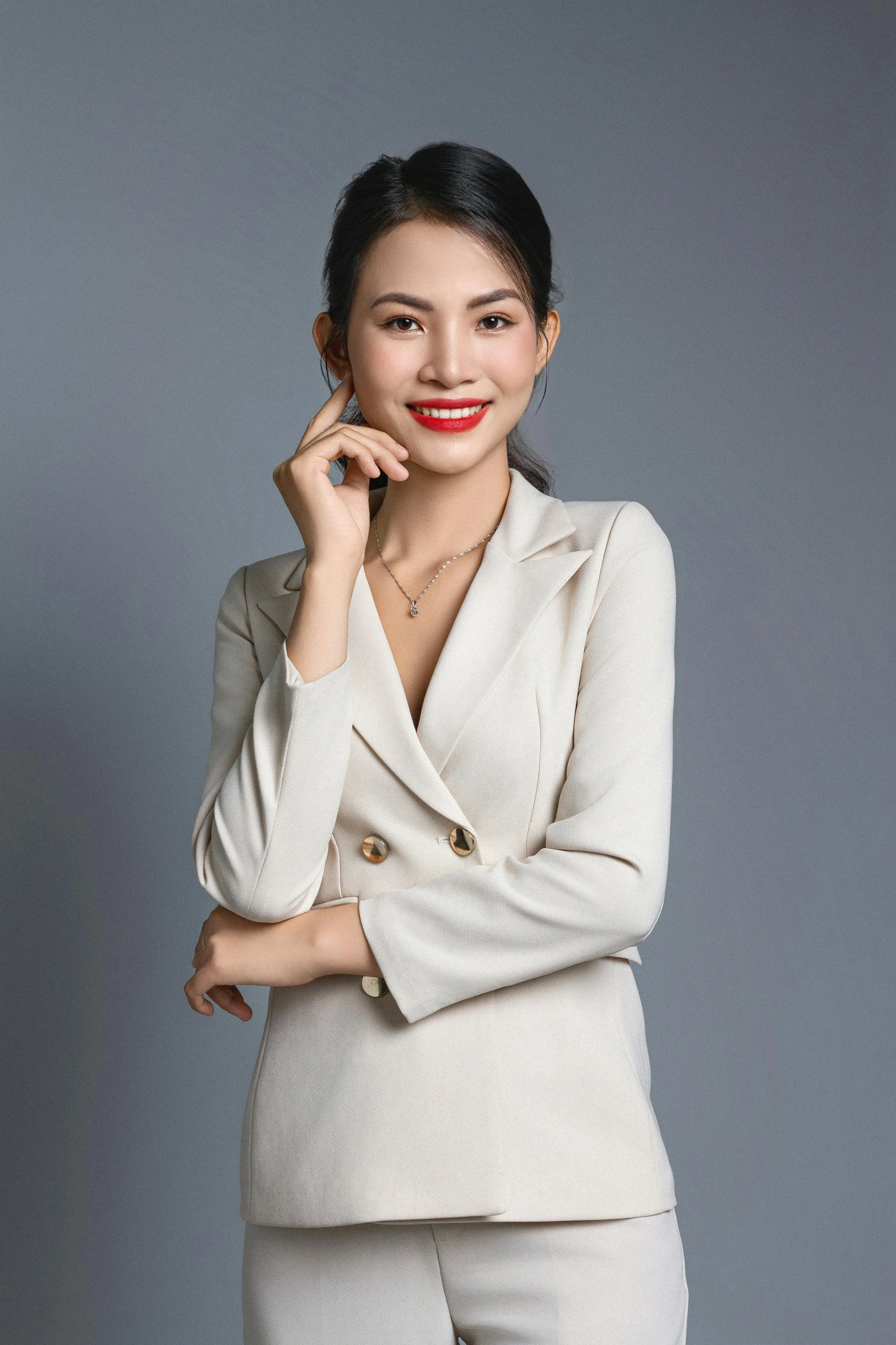 A woman with dark hair, wearing a beige blazer and pants, smiling with red lipstick, and posing with one hand touching her face against a plain grey background.