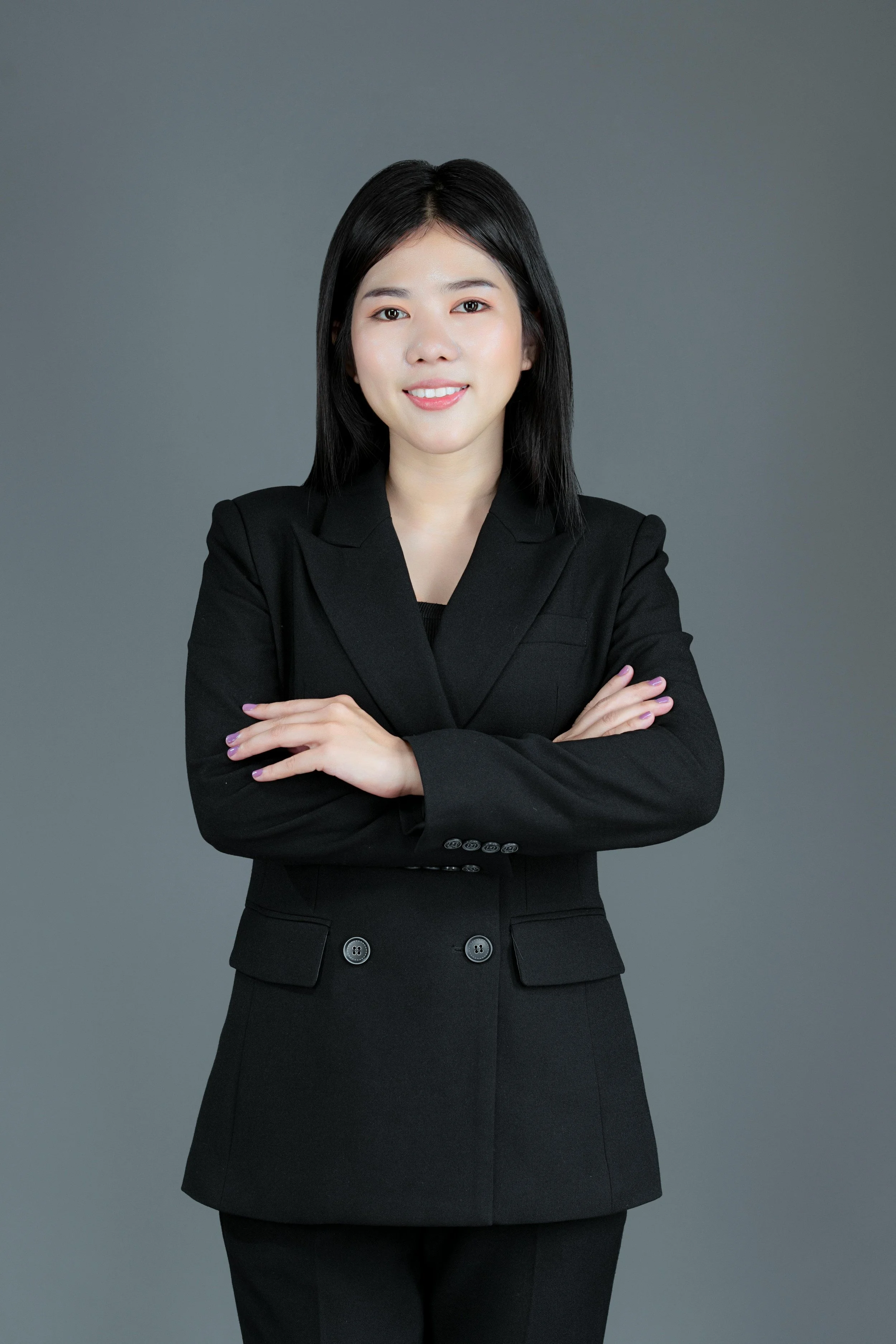 A woman with black hair wearing a black blazer and matching pants, standing with arms crossed against a gray background.