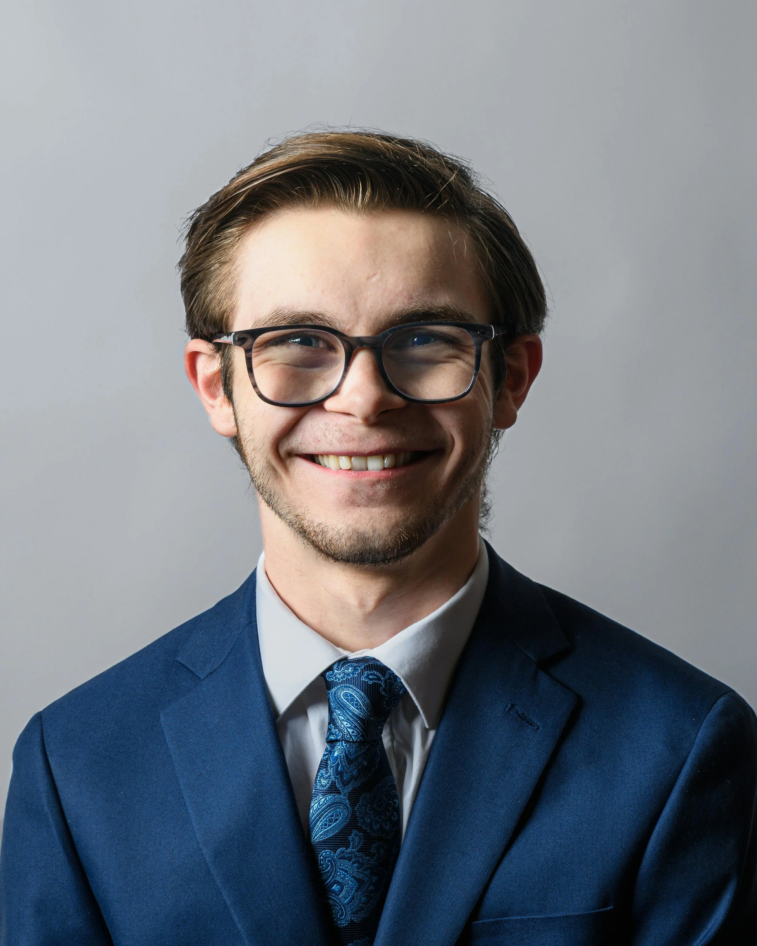A young man with glasses, brown hair, and a beard, wearing a navy suit, white shirt, and a patterned blue tie, smiling against a plain gray background.