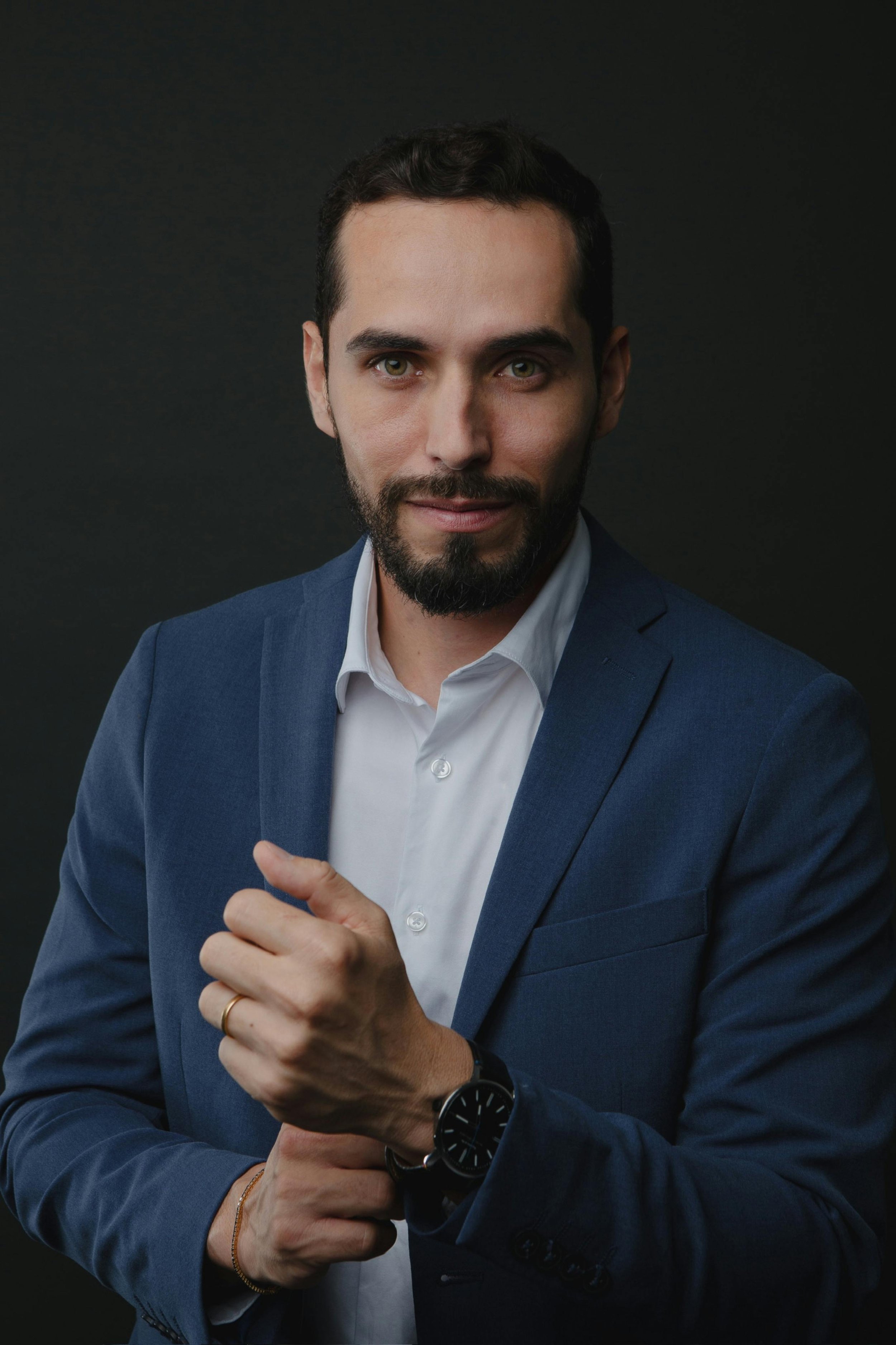 A man in a blue blazer and white shirt pointing at his wristwatch against a black background.