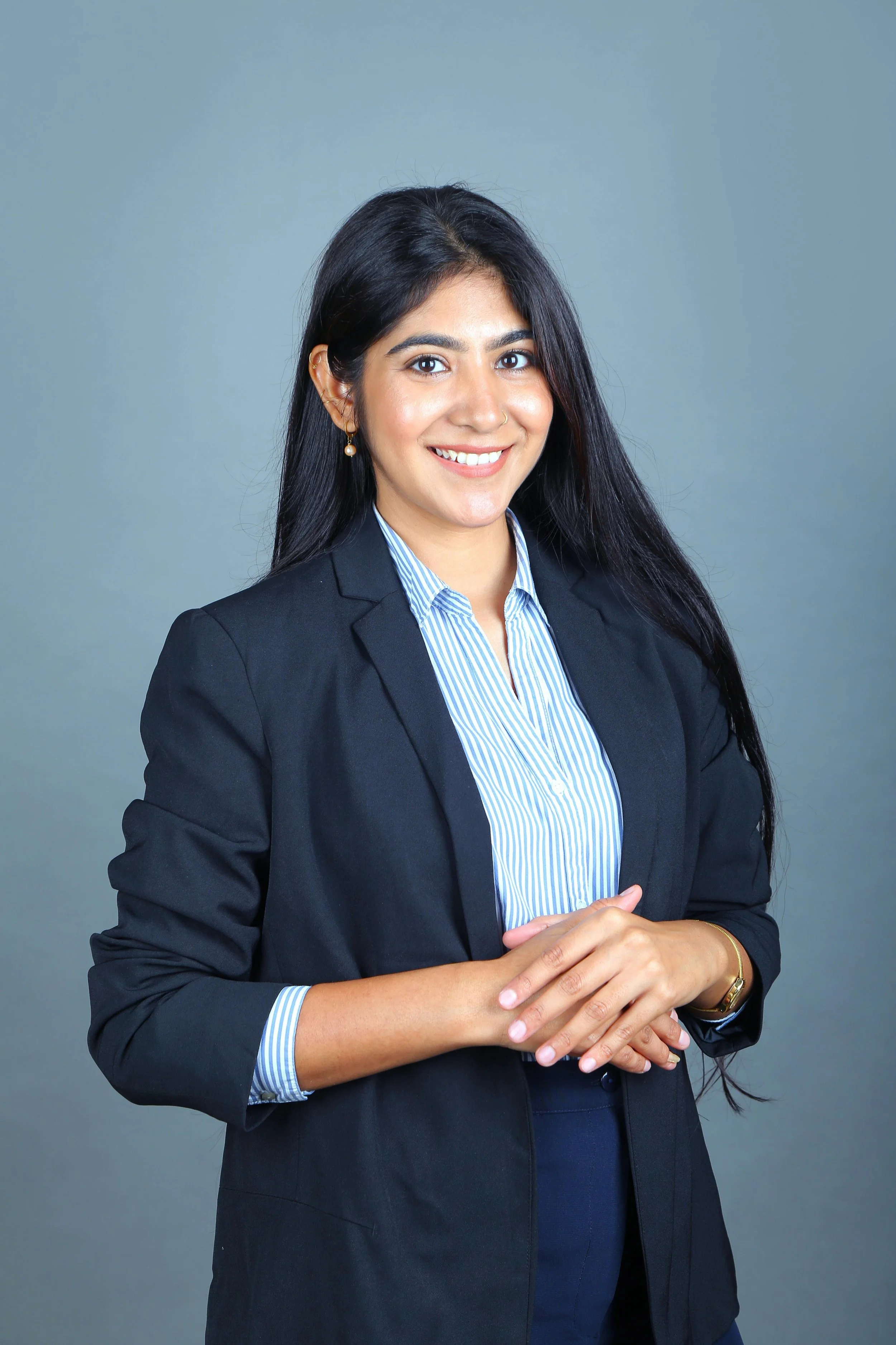 A woman with long black hair, wearing a black blazer over a striped blue and white shirt, standing against a gray background, smiling, with her hands clasped together.