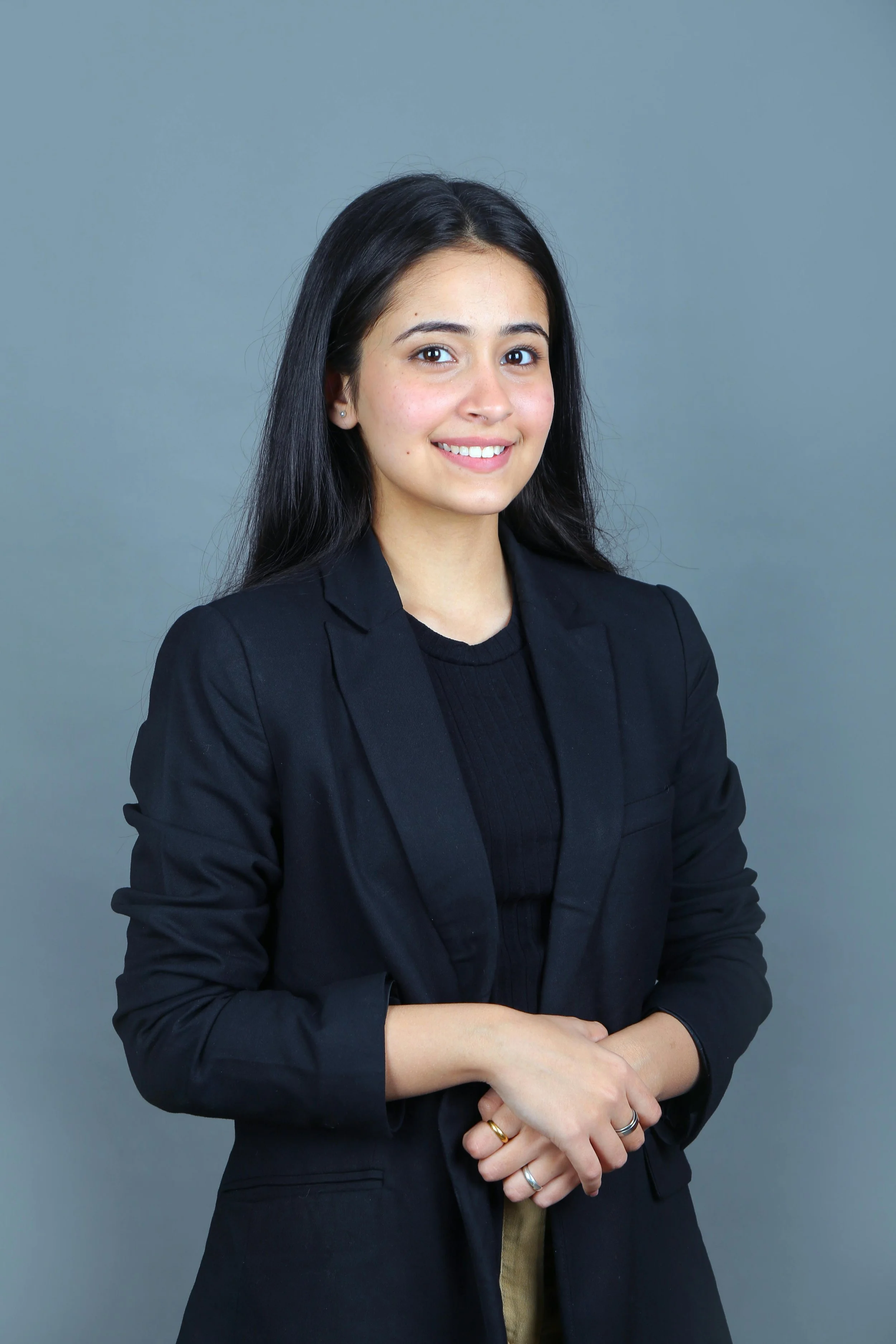 A young woman with long dark hair, wearing a black blazer and smiling, standing against a plain gray background.