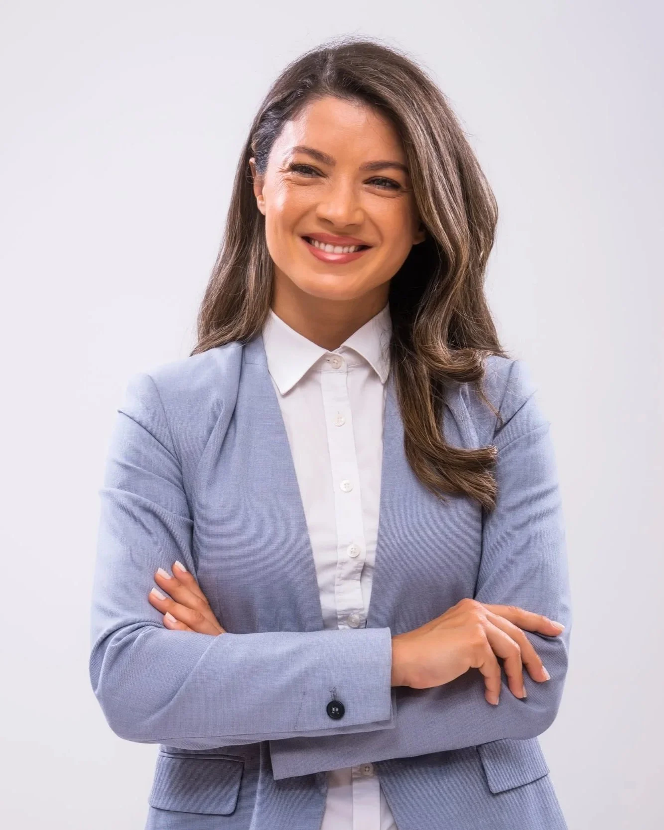 A smiling woman in a light grey blazer and white blouse with crossed arms against a plain background.