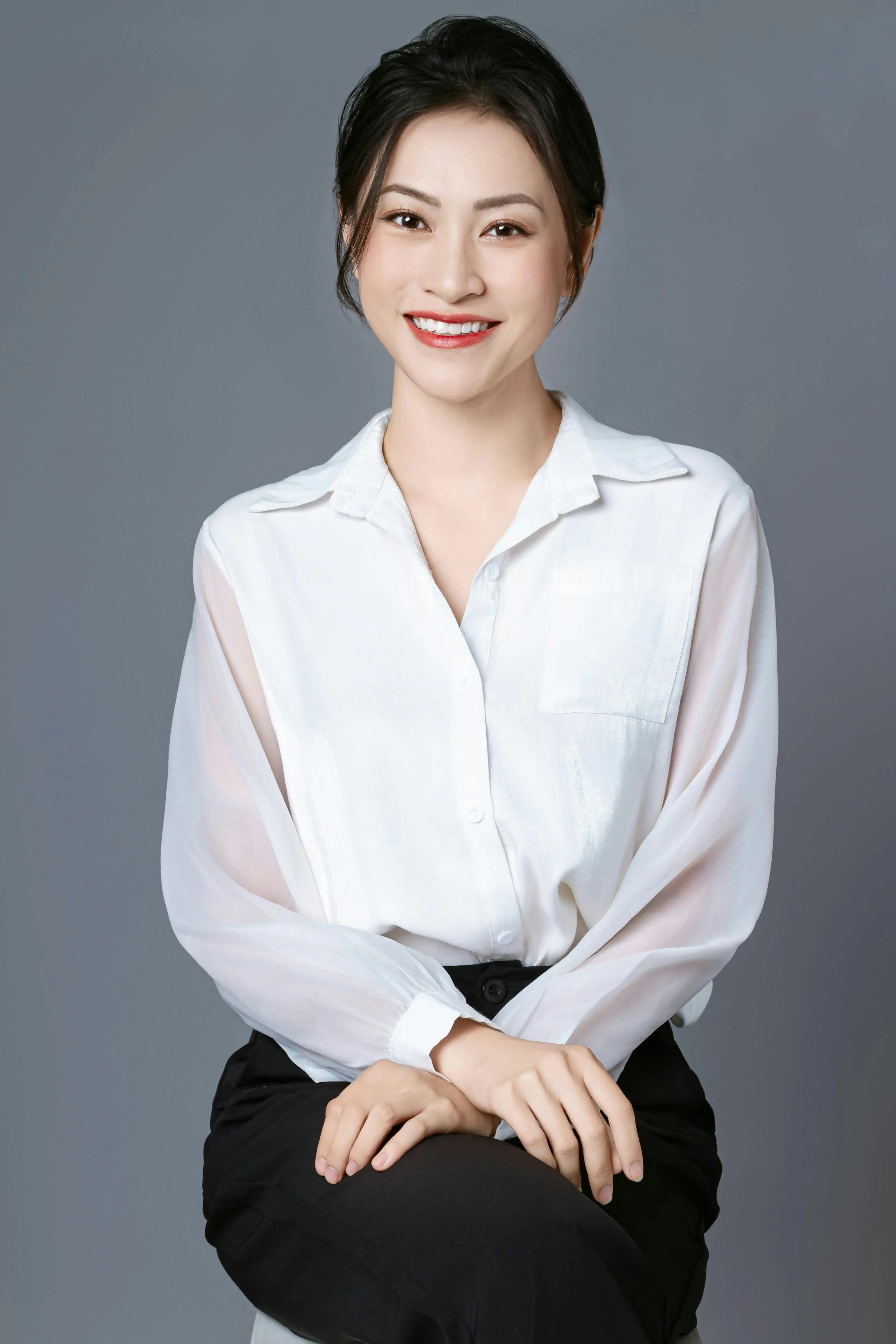 A woman with short dark hair, wearing a white blouse and black pants, smiling while sitting against a gray background.
