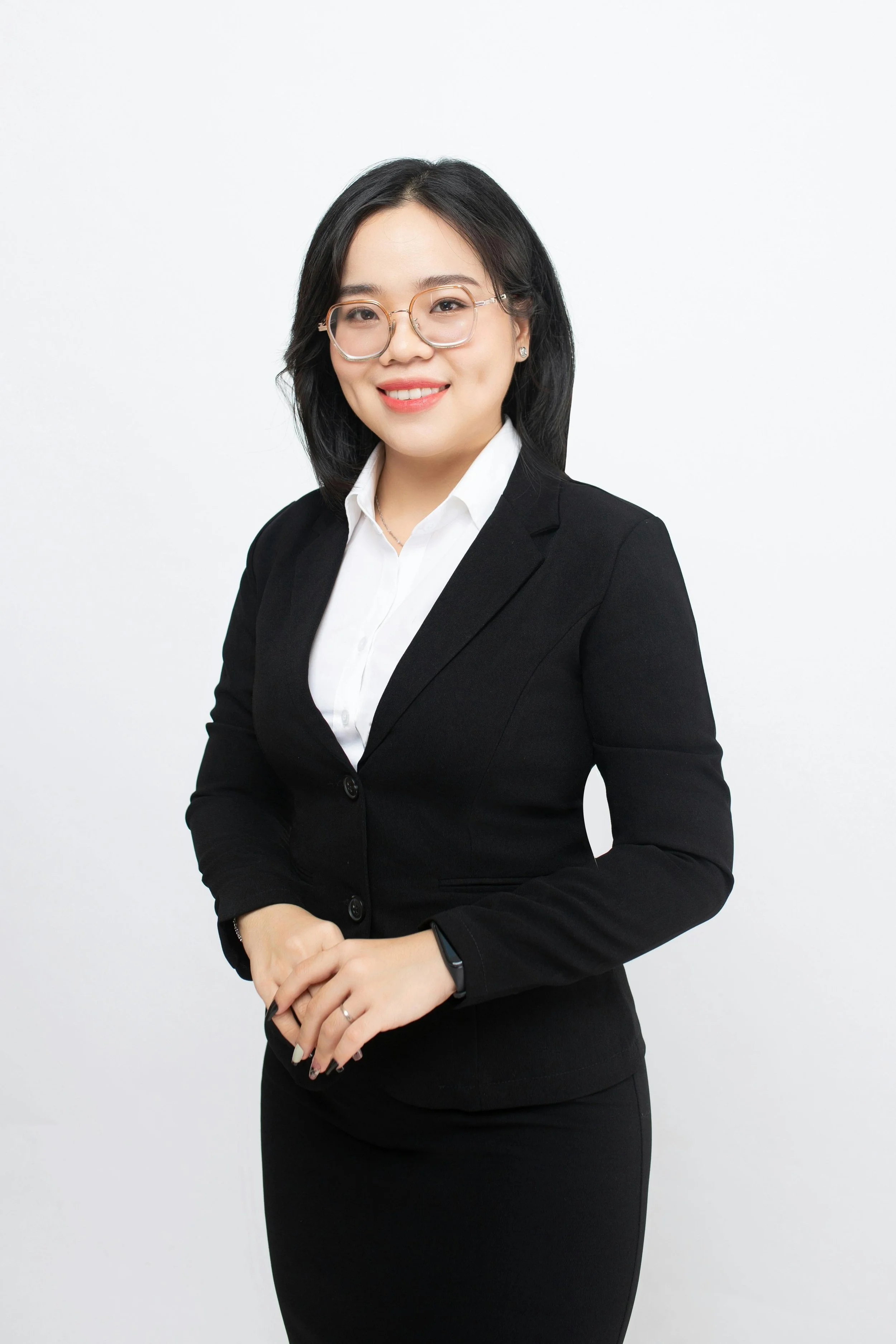 Portrait of a smiling woman with black hair and glasses, wearing a black blazer, white shirt, and black skirt, standing against a plain white background.