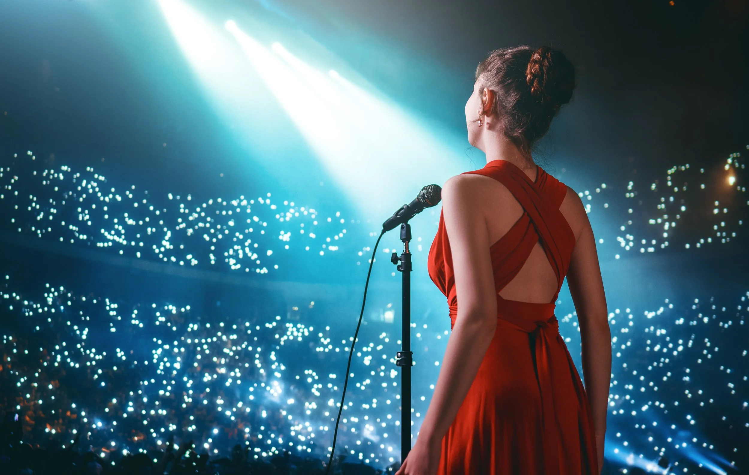 A woman in a red dress stands on stage in front of a microphone, facing a large audience with lights and spotlights creating a sparkling backdrop.