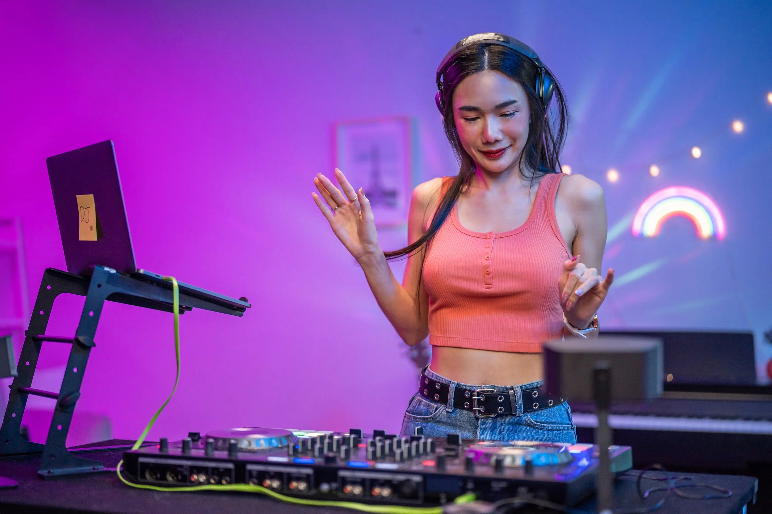Young woman in orange crop top and jeans DJing with headphones in a colorful neon-lit room.
