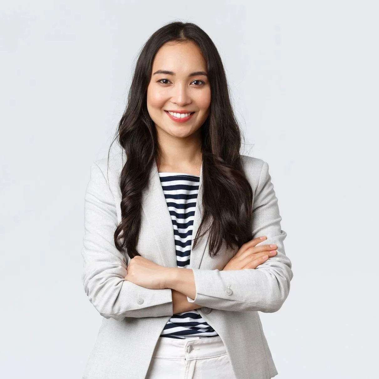 A young woman with long dark hair, smiling, wearing a light-colored blazer over a striped shirt, with arms crossed, standing against a light gray background.