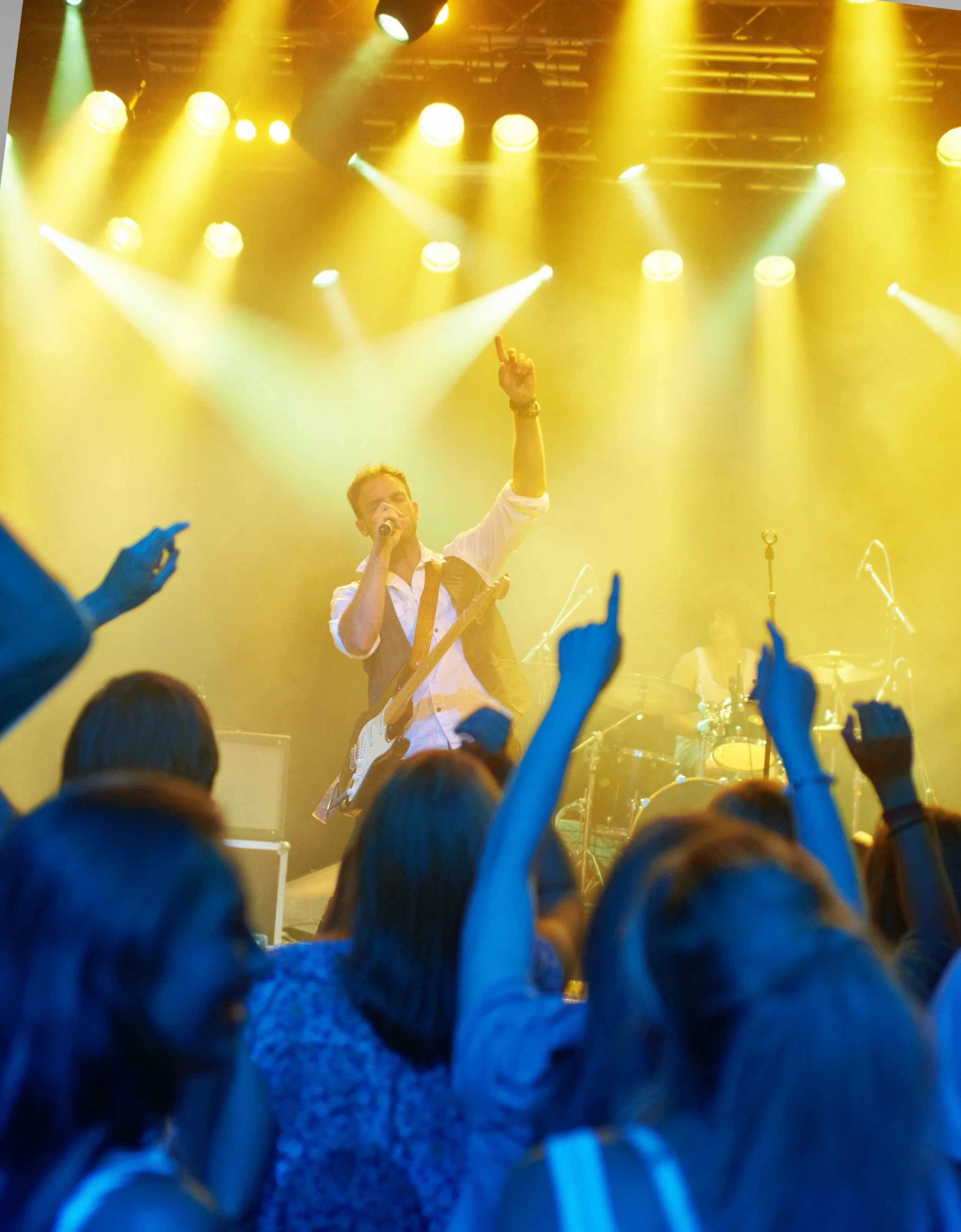 A musician on stage performing at a concert with yellow stage lights, surrounded by an enthusiastic crowd.