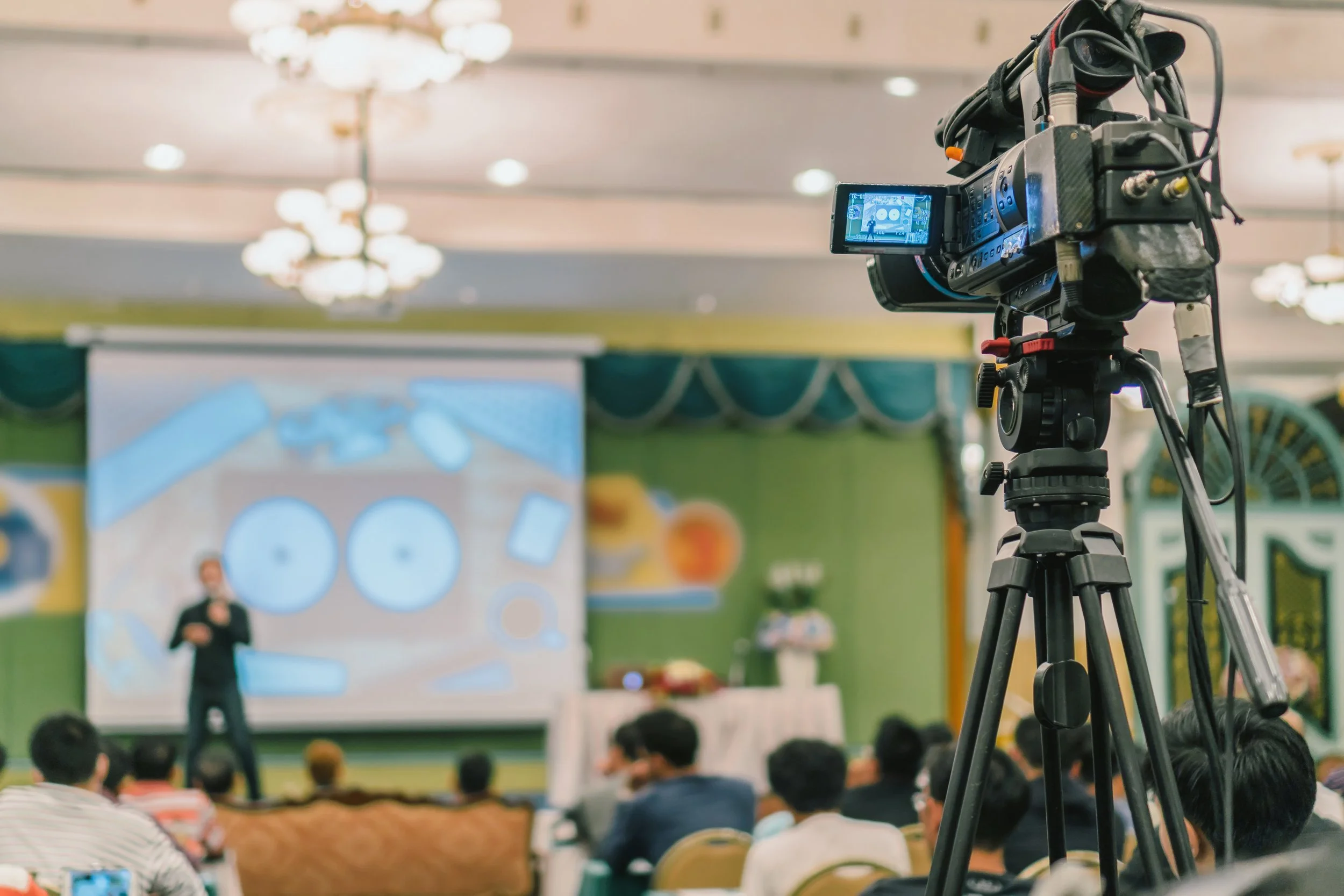 Video camera on tripod recording a speaker at a conference, with audience seated facing a presentation screen in a decorated hall.