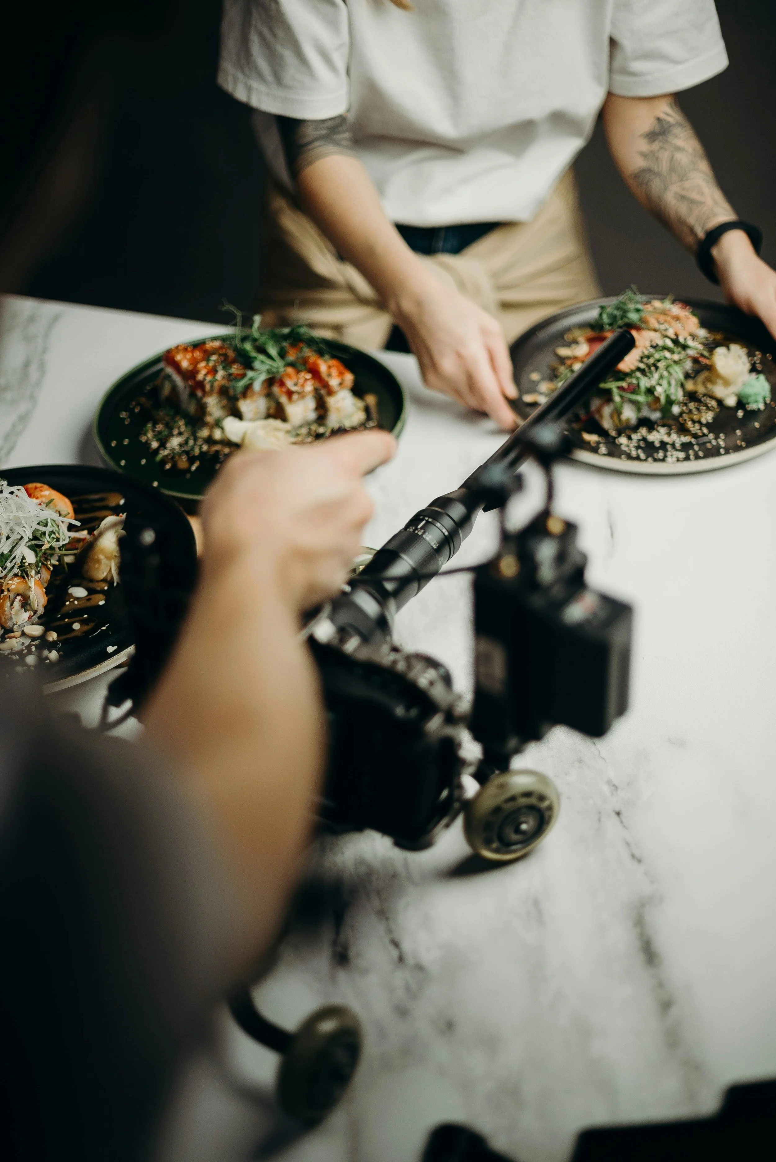 Person filming sushi dishes on black plates at a white table.